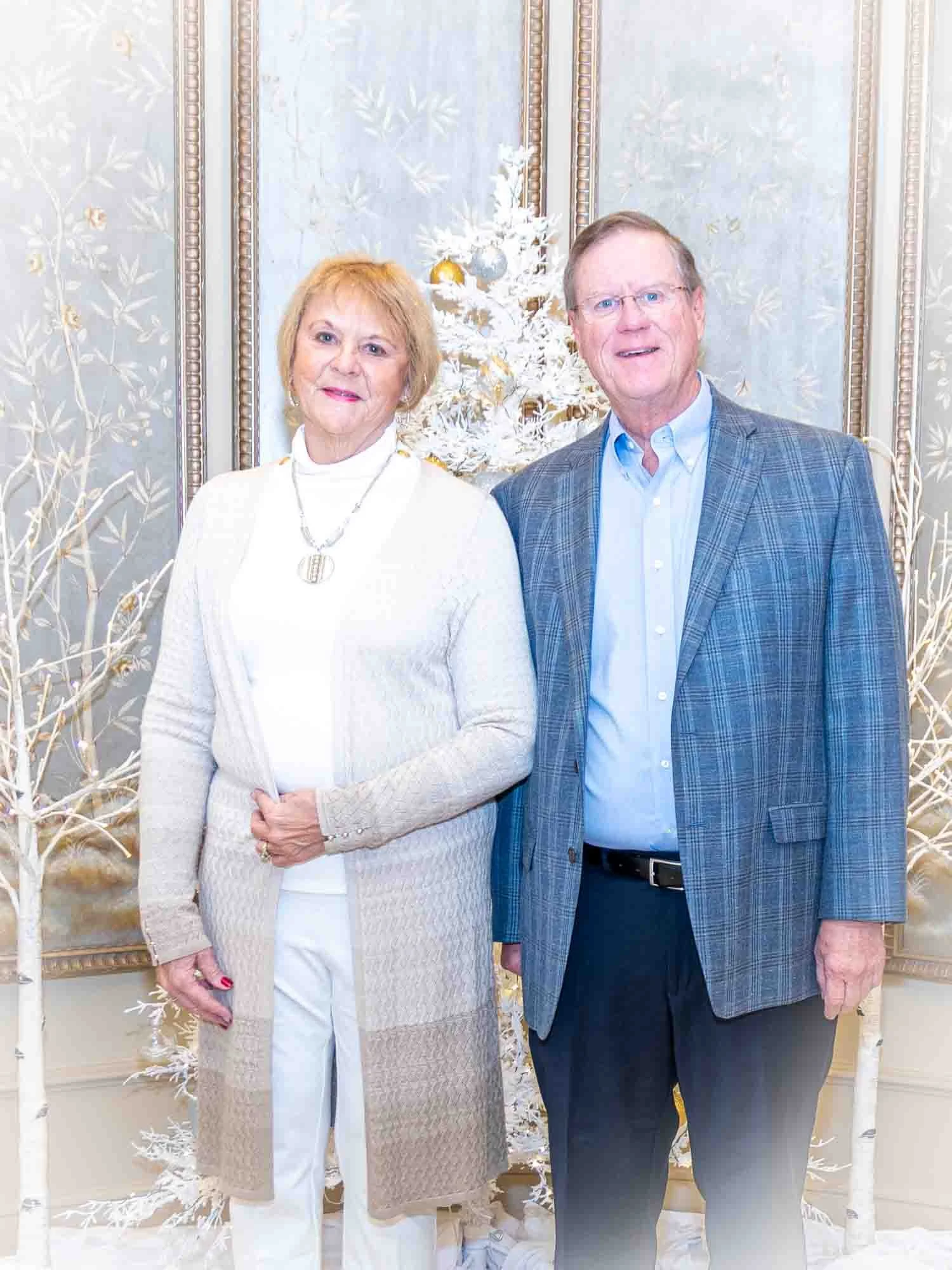 A couple standing in front of a decorated white Christmas tree indoors; the man is wearing a blue checkered blazer and the woman is wearing a beige cardigan.