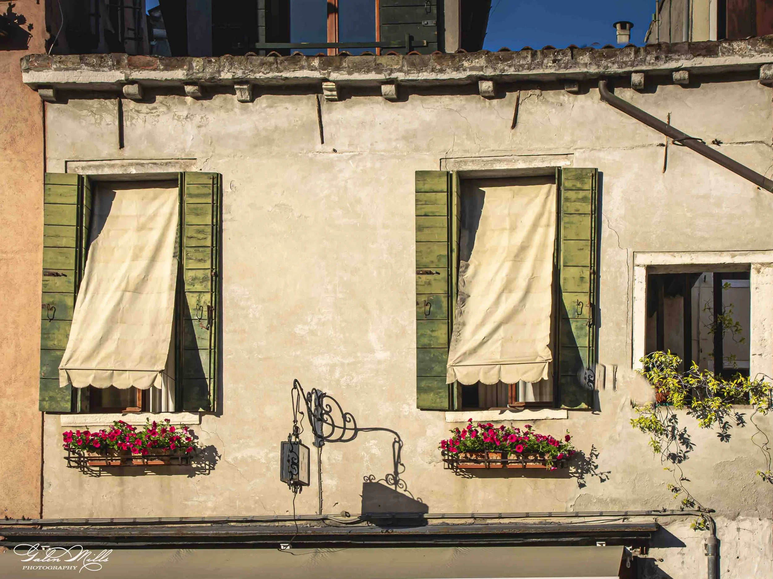 The image depicts the upper portion of a rustic building with two windows featuring green shutters and beige awnings. There are flower boxes with pink flowers under each window and a small black iron sign hanging between the windows. The wall appears