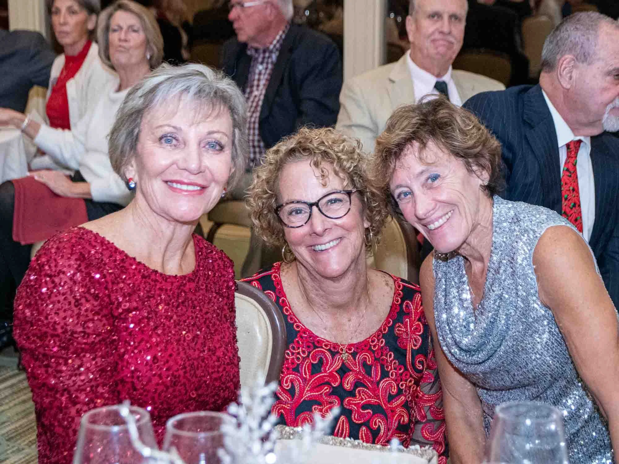Three women smiling at a formal event, wearing sequined dresses, with people seated in the background.