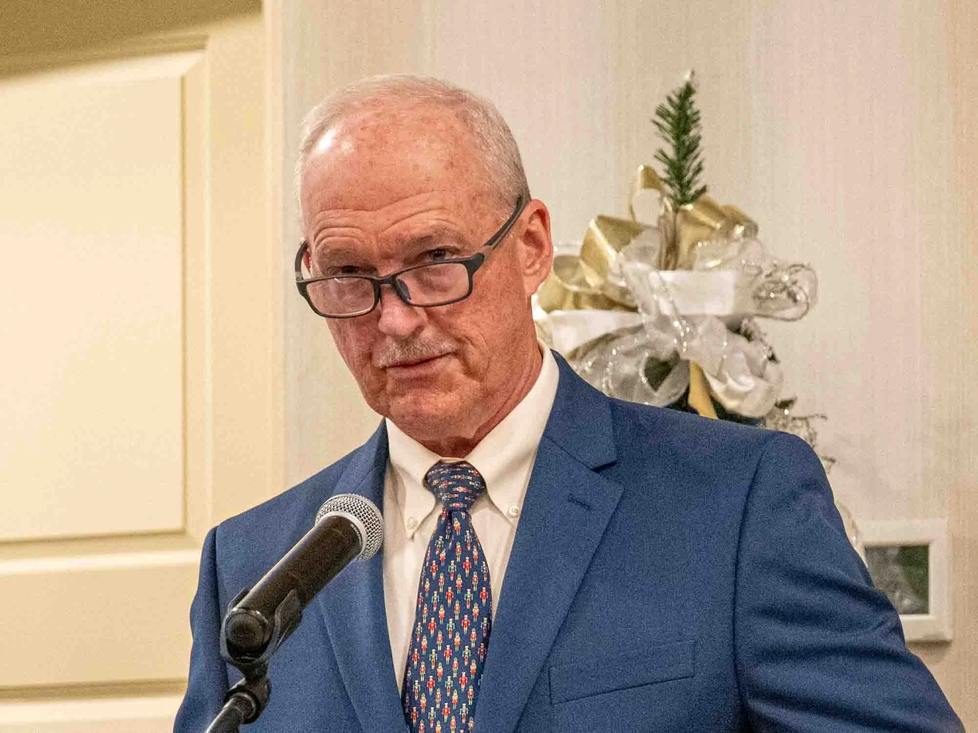 A man in a blue suit and glasses speaking at a microphone indoors.