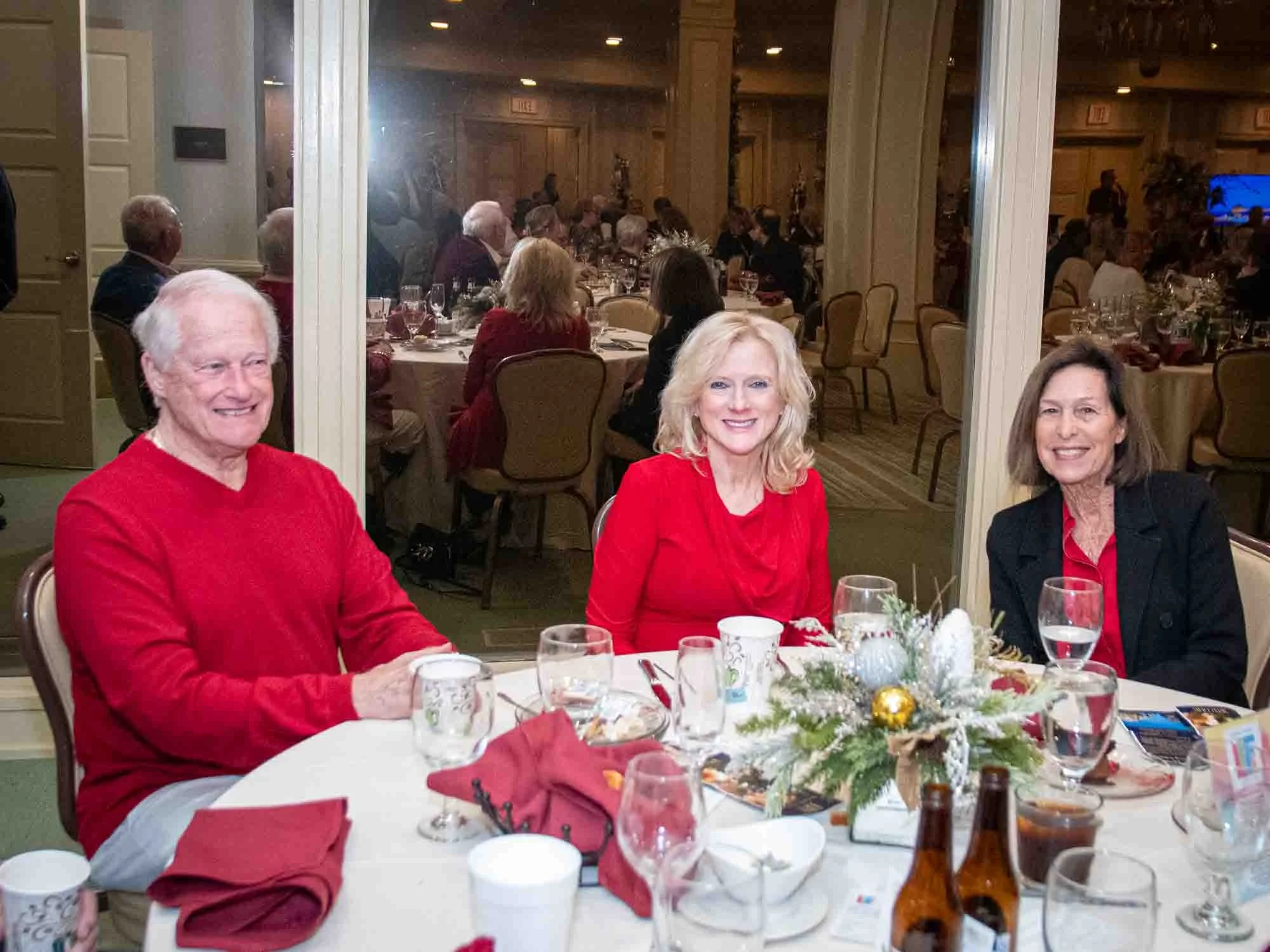 Three people sitting at a round dining table during a festive event. The table is set with drinks, glasses, and a centerpiece with greenery and ornaments. The people are smiling and wearing red, possibly indicating a holiday season gathering. Other t