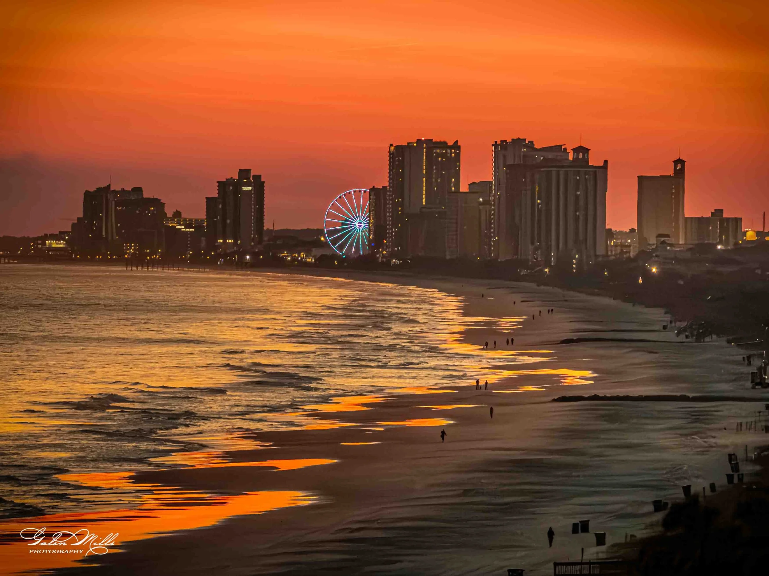 A city skyline at sunset with tall buildings, a Ferris wheel, and the ocean in the foreground, reflecting the orange sky.