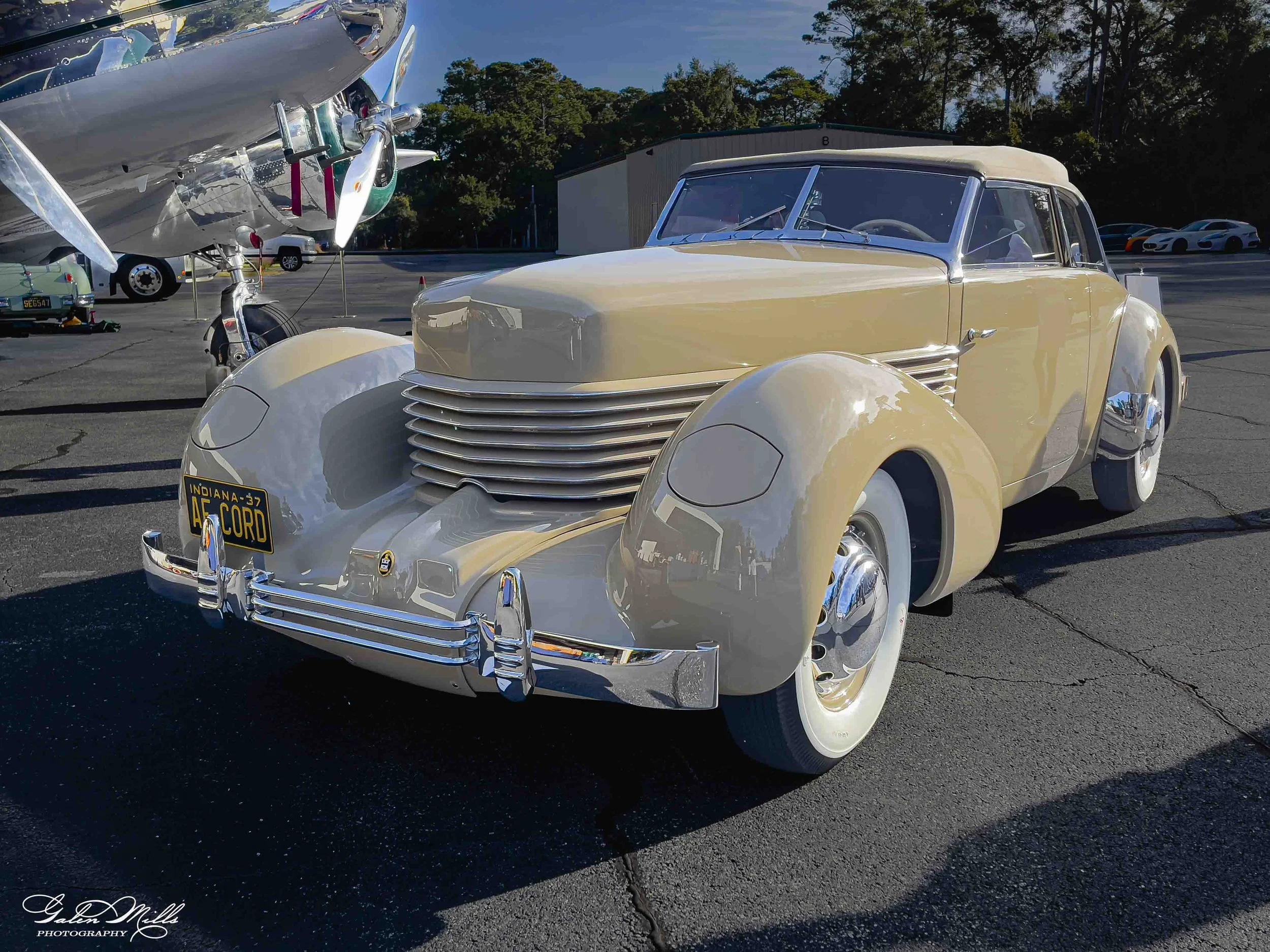 Vintage cream-colored car with chrome accents parked at an outdoor event, with several modern cars and a hangar in the background, under a clear blue sky.