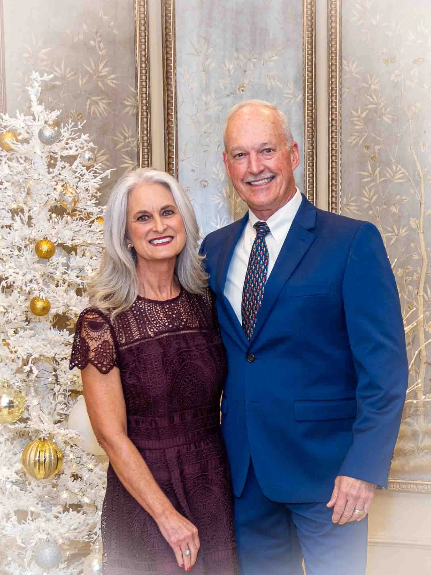 A mature couple posing indoors in front of a decorated white Christmas tree. The woman is wearing a dark purple lace dress, and the man is in a blue suit with a multicolored tie. The background features ornate decorative panels.