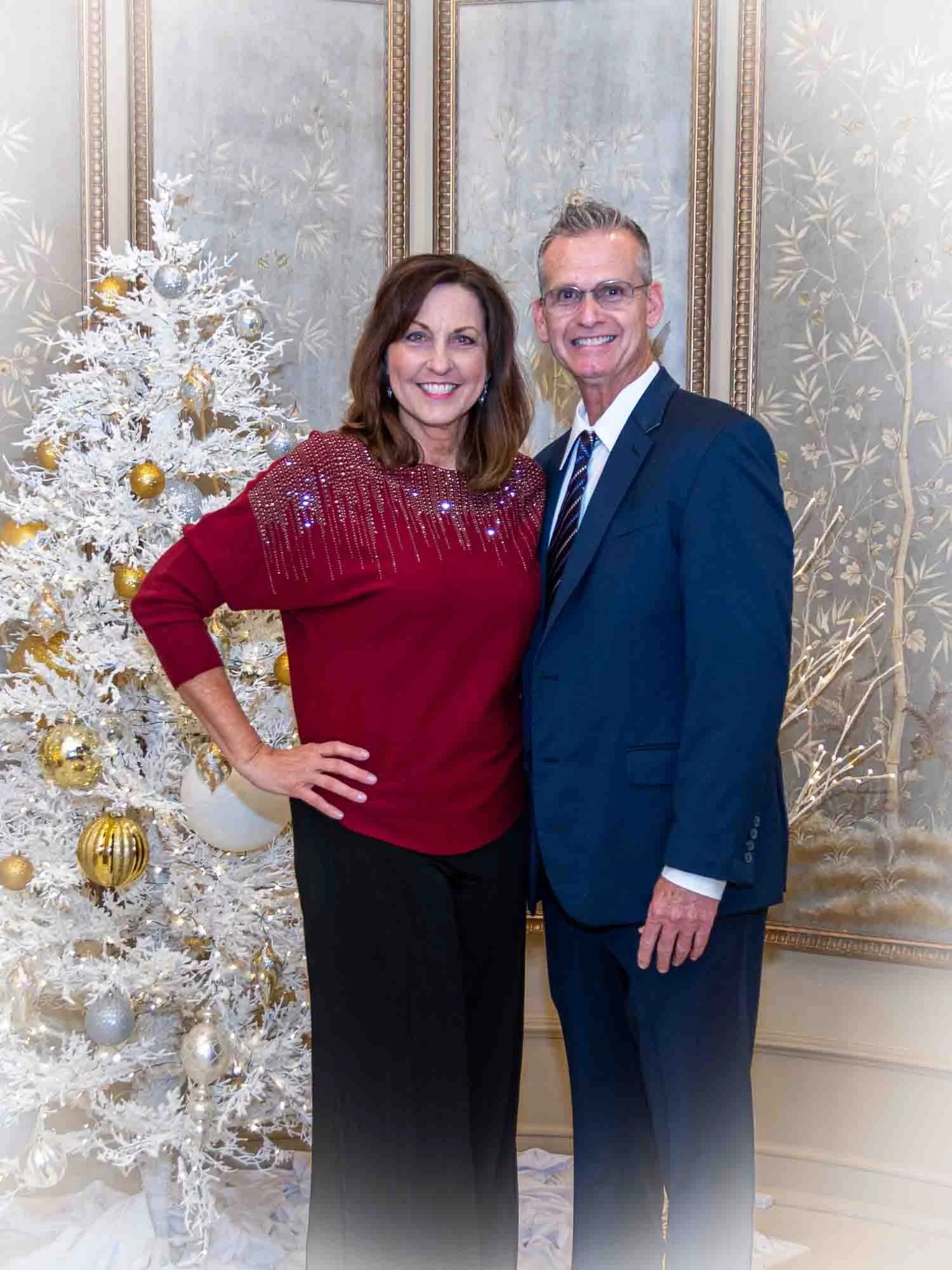 A man and a woman smiling in front of a decorated white Christmas tree with gold and silver ornaments, standing in a sophisticated room with ornate paneling.