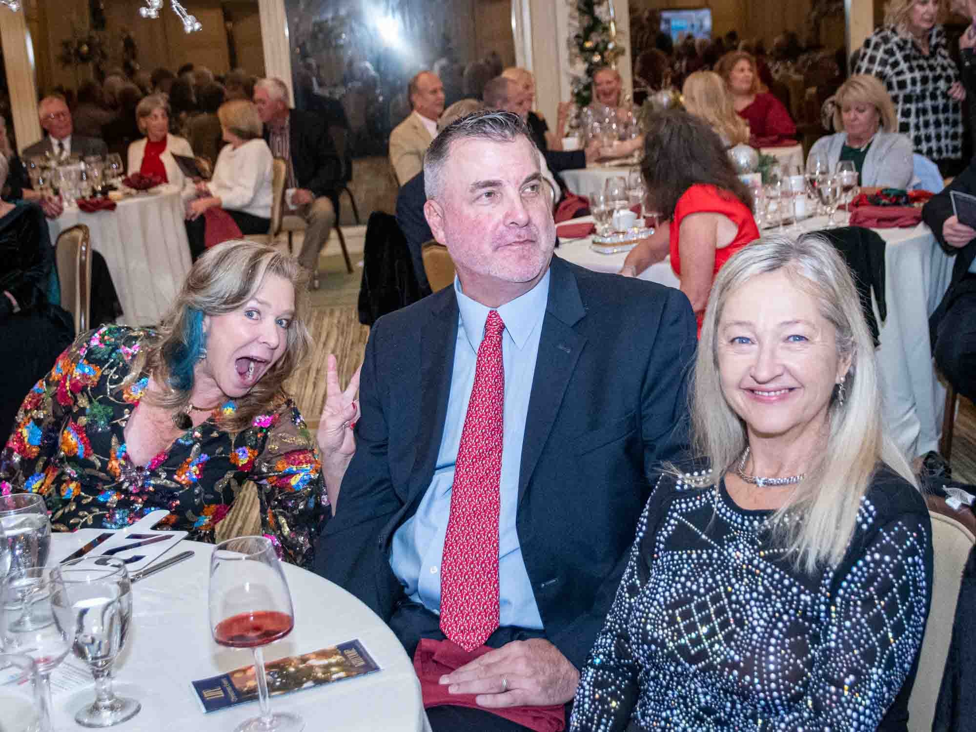 Group of people at a formal dinner event, with one woman playfully posing with a peace sign. Tables are set with drinks and decorations.