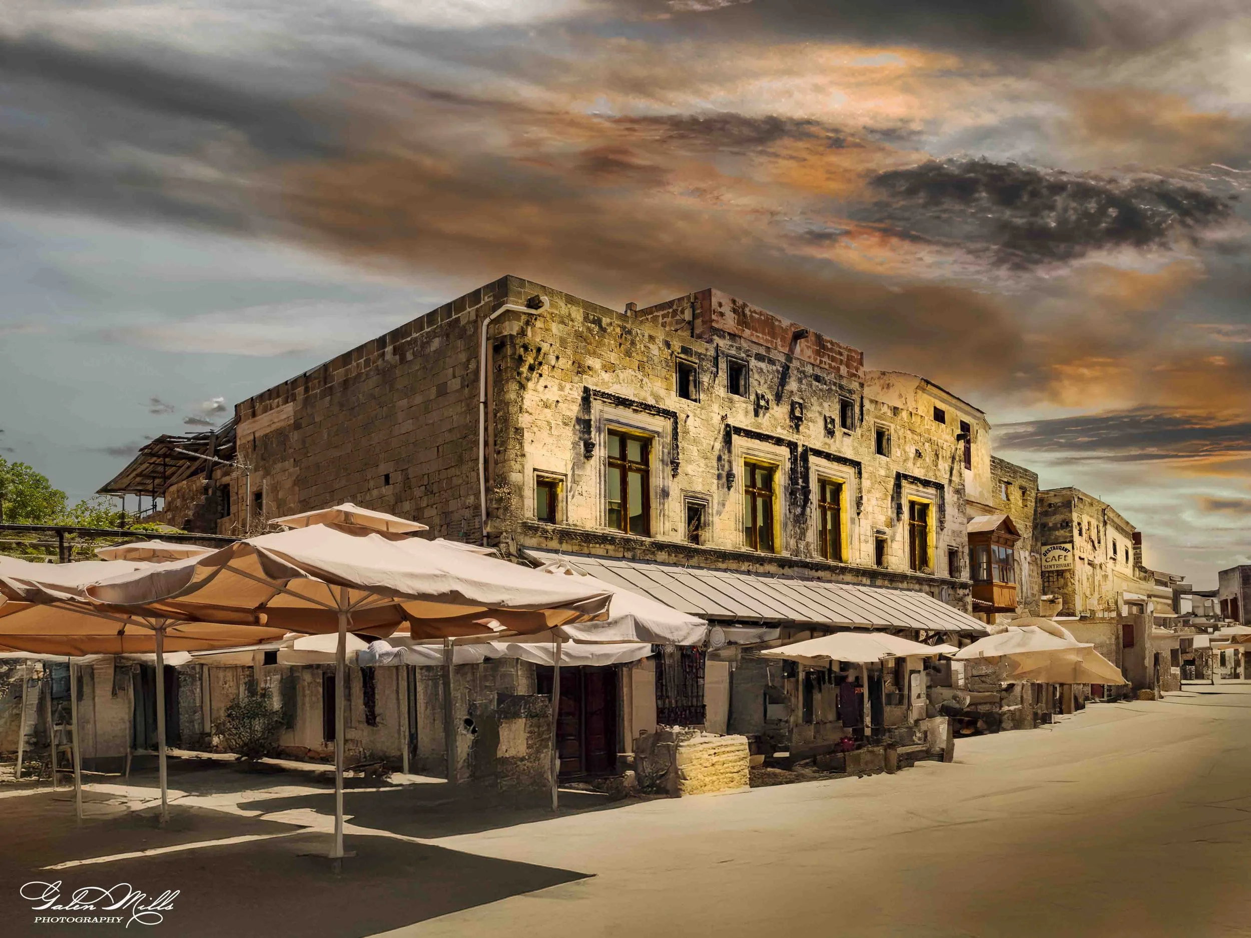 Historic stone building with outdoor café tables and umbrellas under a dramatic sky.