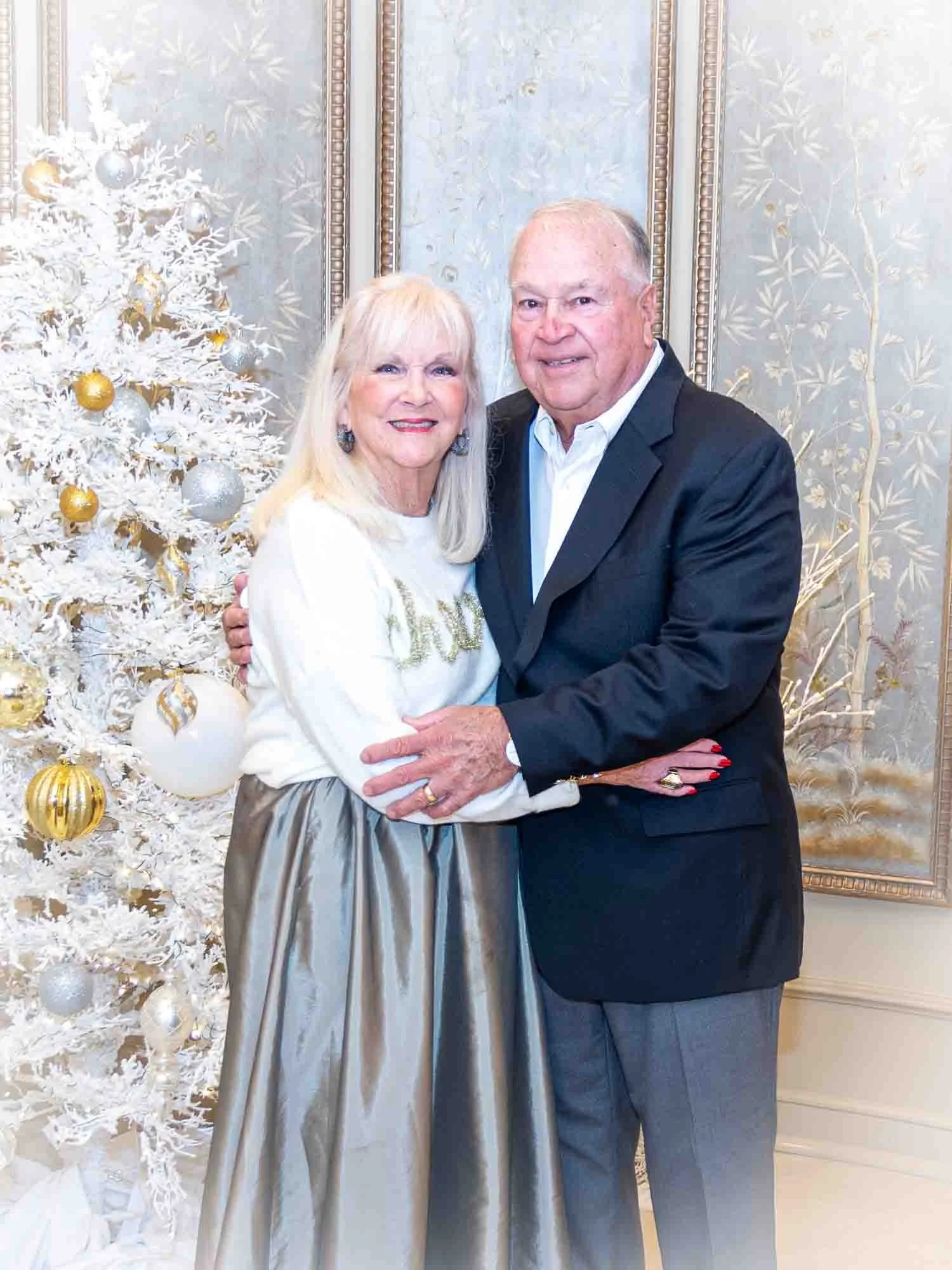 An elderly couple standing in front of a white Christmas tree decorated with gold and silver ornaments, embracing each other. The woman is wearing a white sweater and a long silver skirt, while the man is in a formal dark suit. The background feature