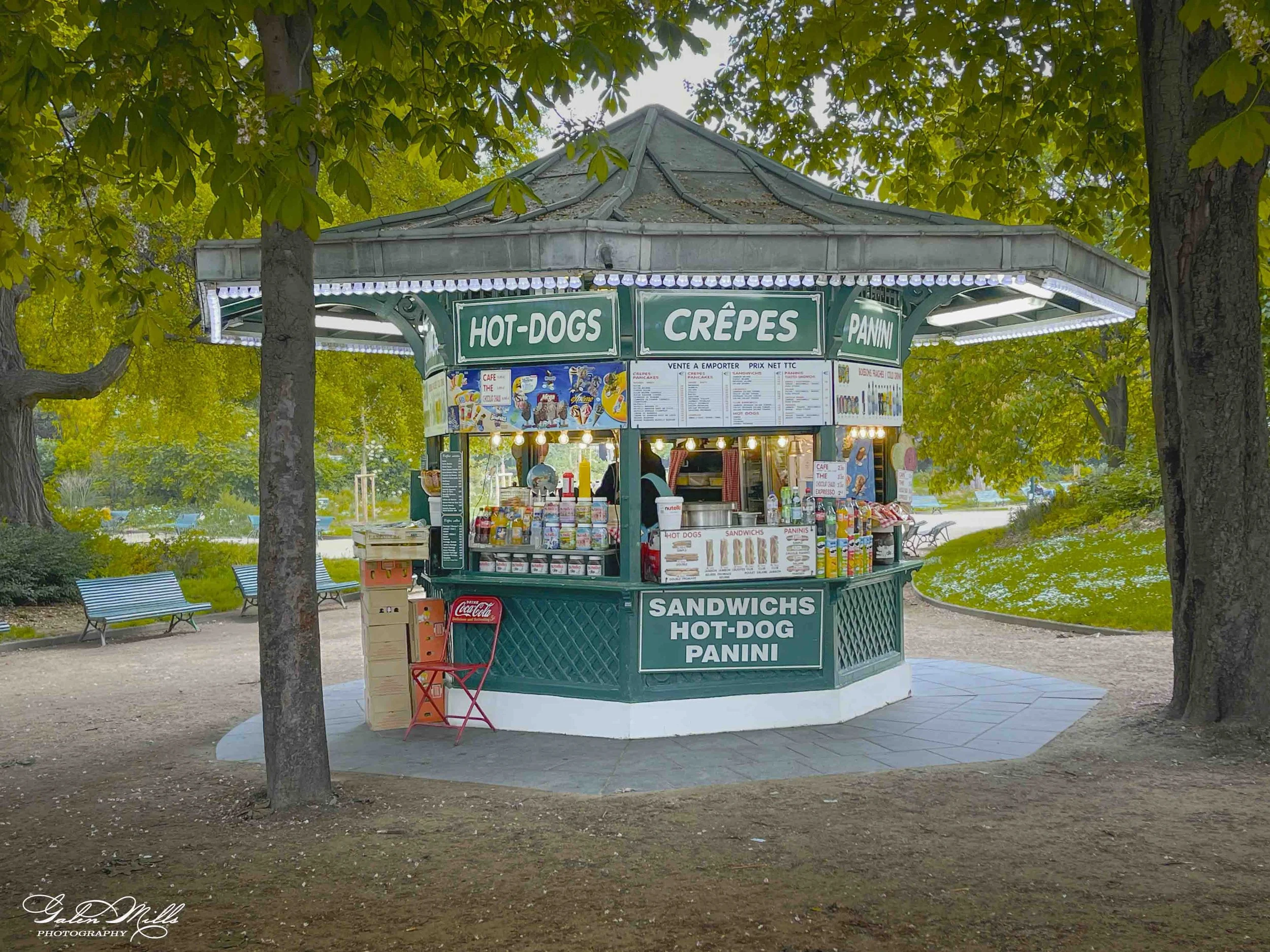 A small green kiosk in a park surrounded by trees, selling snacks like hot dogs, crepes, paninis, and sandwiches, with menu signs and various condiments visible.