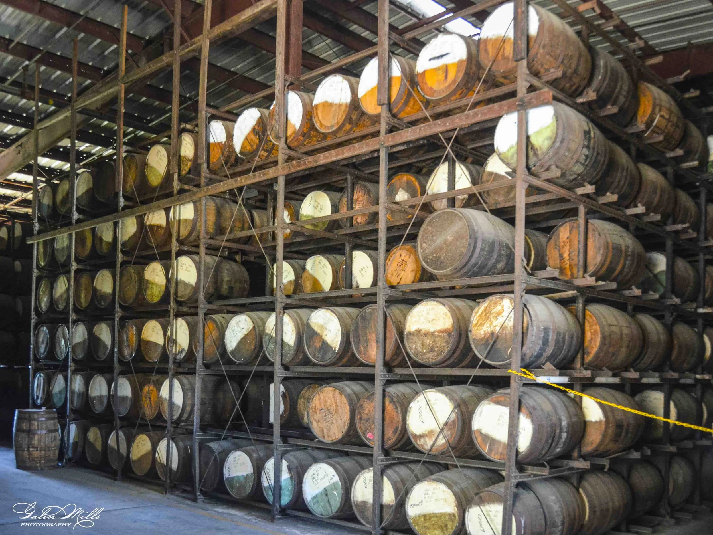 Tall metal storage racks filled with stacked wooden barrels, some marked with white and orange paint, inside a warehouse or distillery.