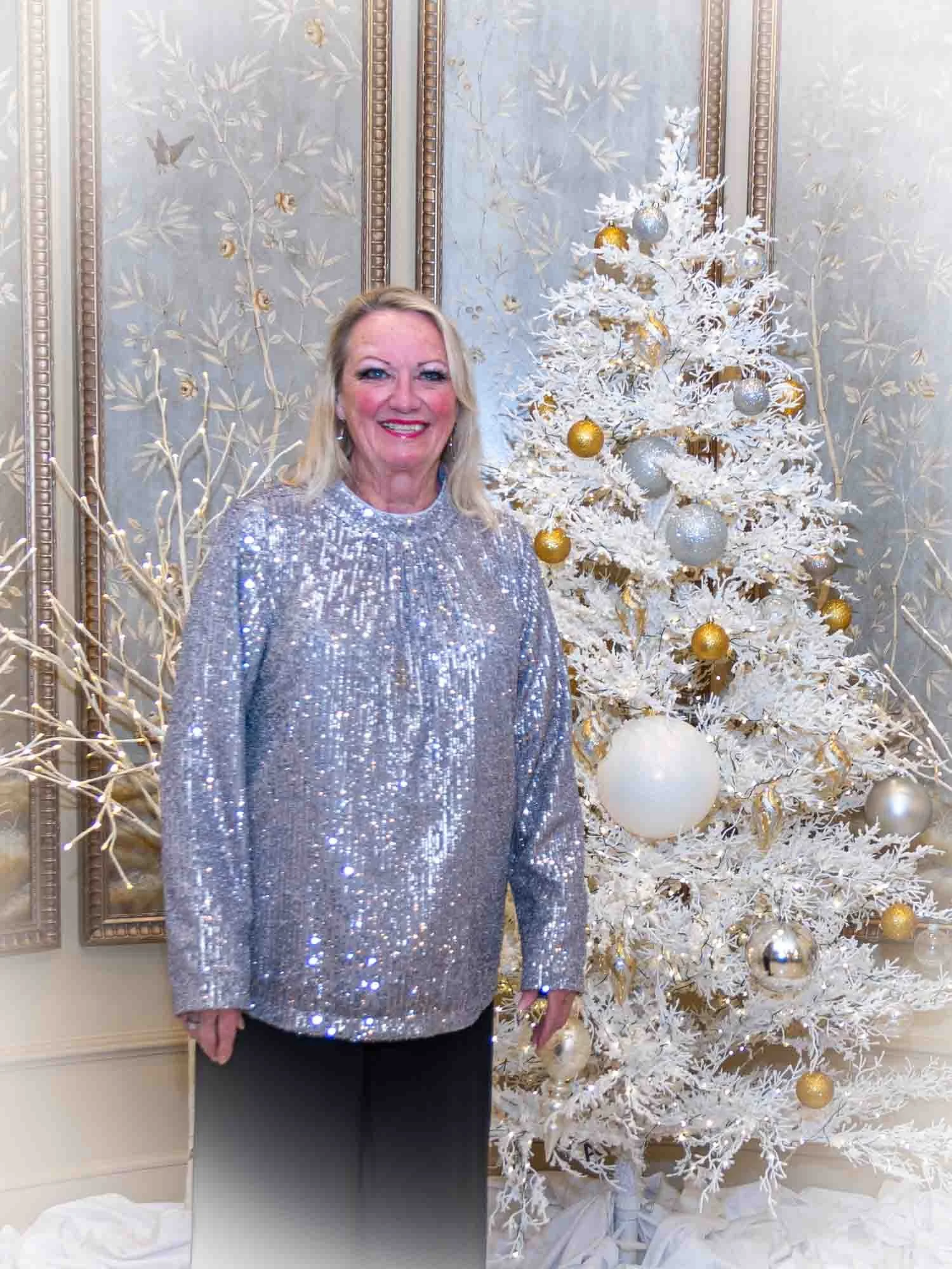 Woman standing in front of a decorated white Christmas tree wearing a silver sequined top.