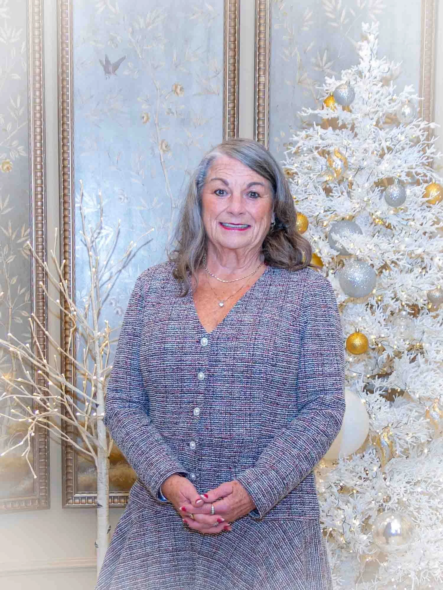 Smiling older woman standing in front of a white Christmas tree with gold and silver ornaments, in a decorated room with framed art on the wall.