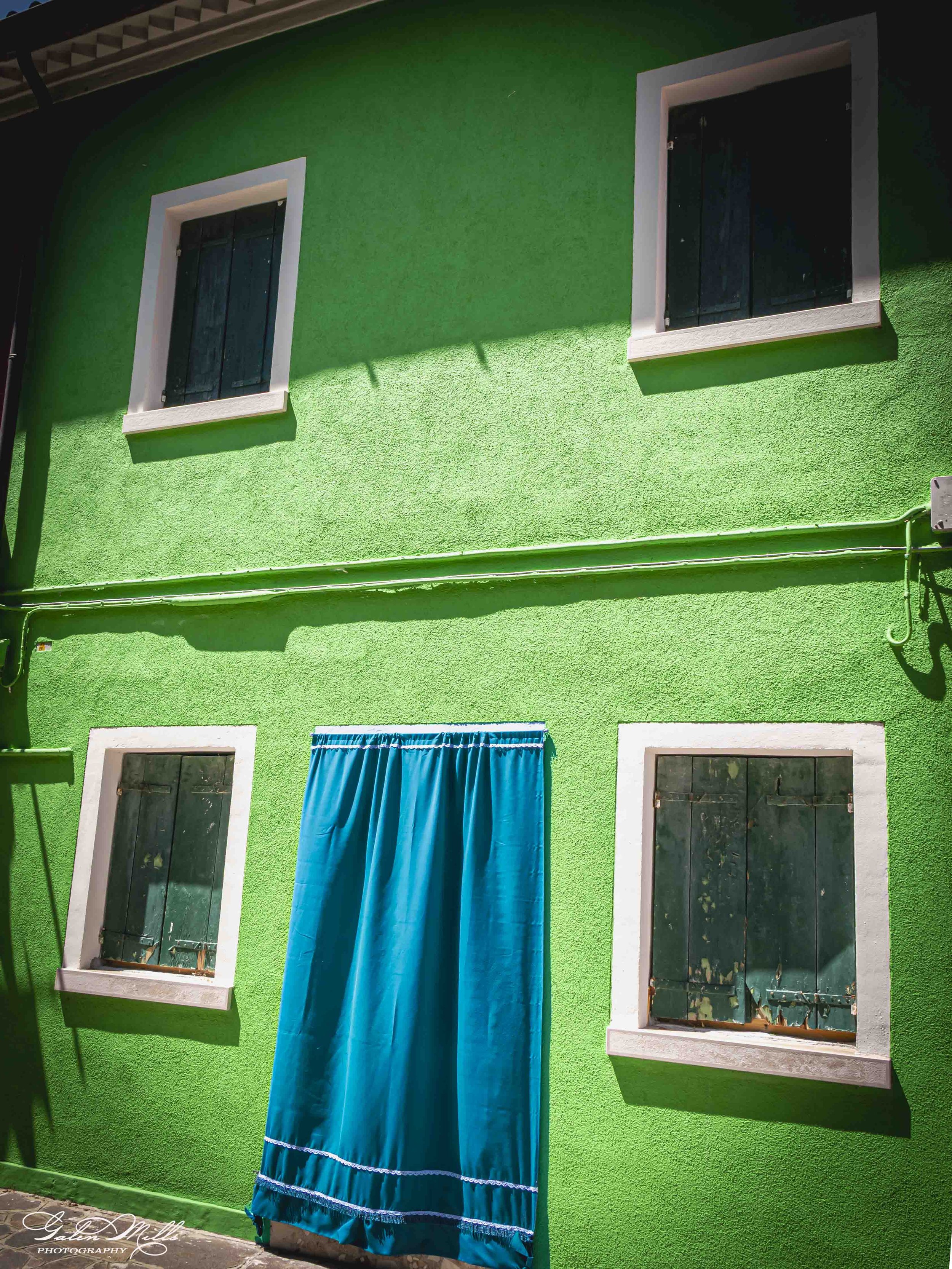 Bright green house with three windows with black shutters and one blue curtain covering a window. The house has a white trim around the windows.