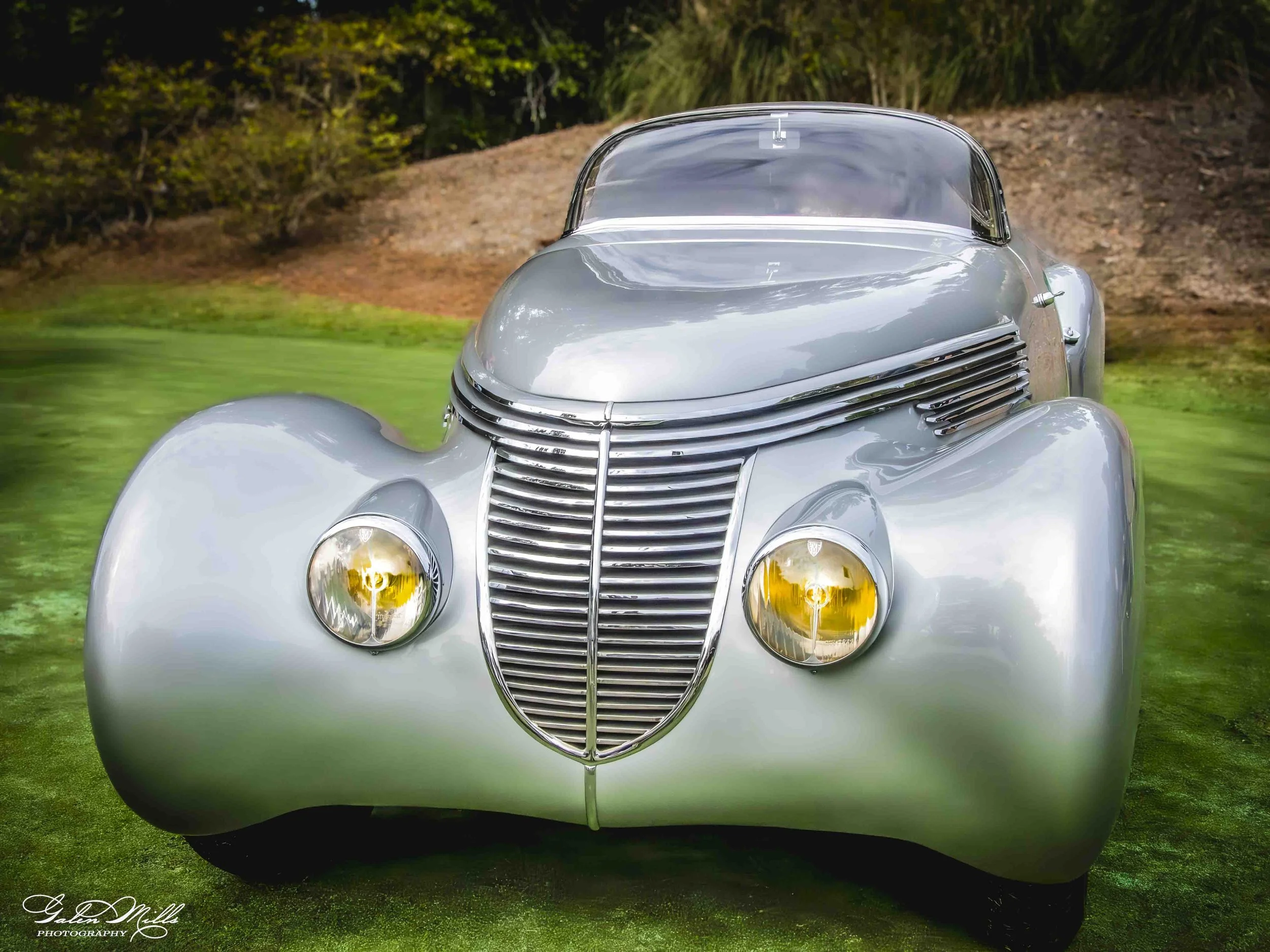 Front view of a vintage silver car with a rounded body, large grille, and distinctive headlights, parked on grass with trees in the background.