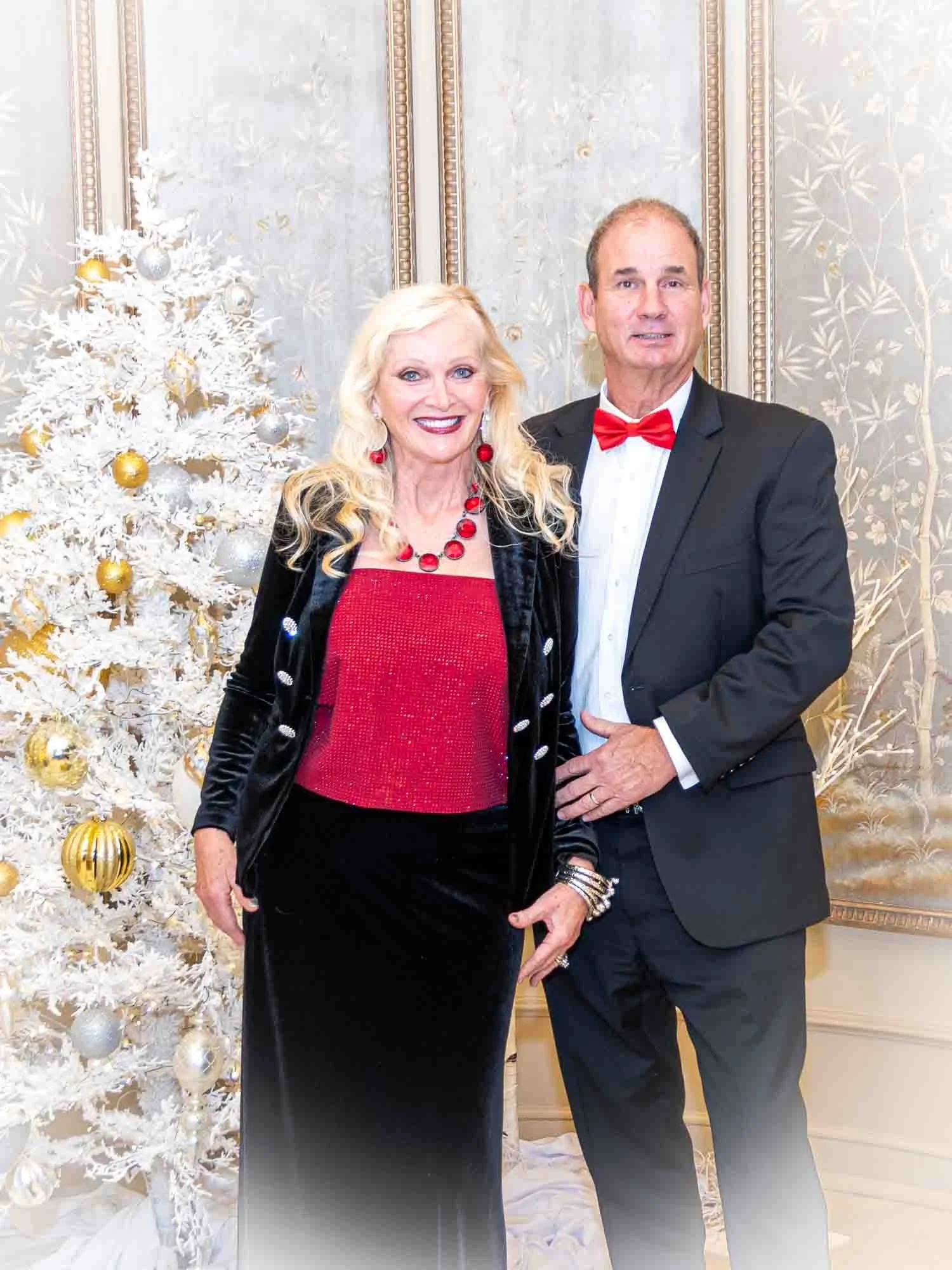 A well-dressed man and woman posing in front of a white and gold Christmas tree.