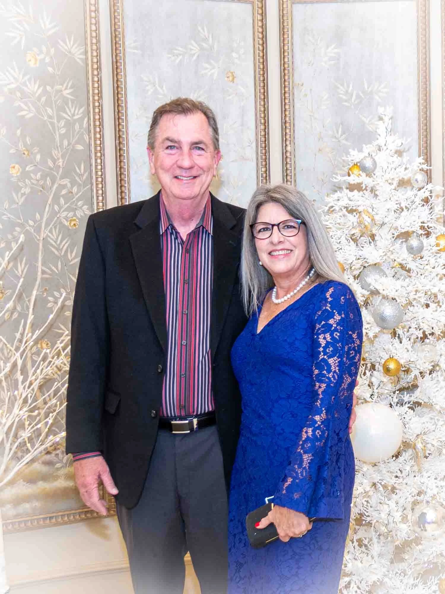 A smiling couple posing beside a decorated white Christmas tree indoors. The man is wearing a black suit jacket and a striped shirt, while the woman is in a blue lace dress. The background features ornate paneling with a subtle floral design.
