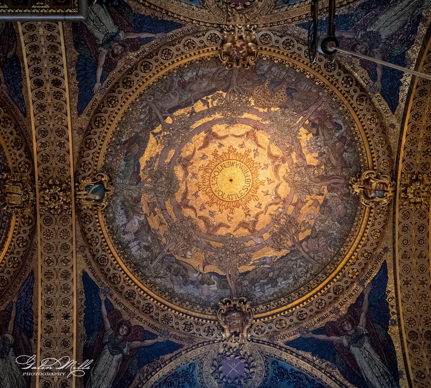 Ornate ceiling with golden mosaic and decorative patterns, including Latin inscriptions and angelic figures, in a circular design.