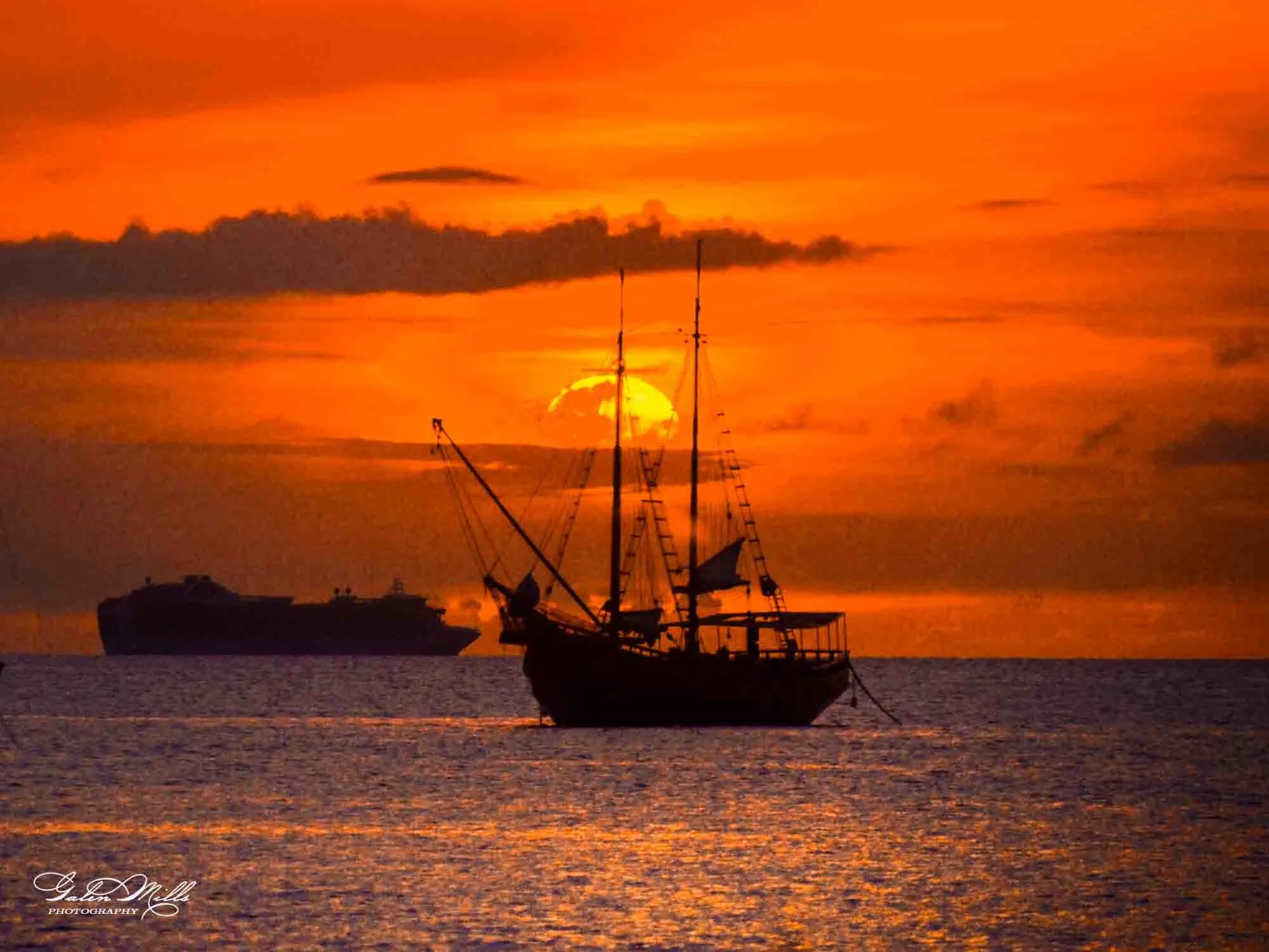 Silhouette of a sailing ship at sea during sunset with a large ship in the background and orange sky.