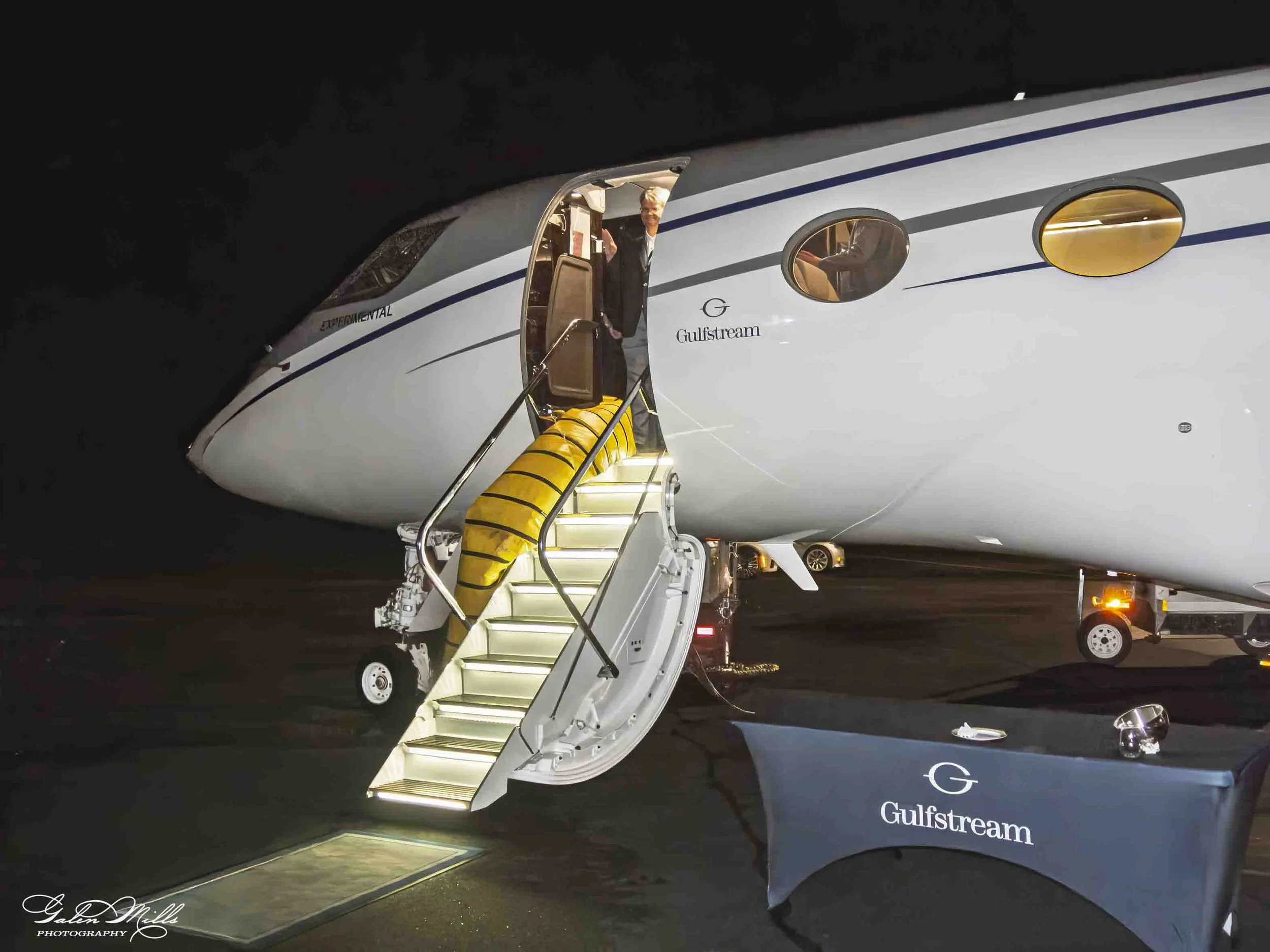 A Gulfstream private jet at night with a set of stairs leading up to its entrance. An individual is visible at the top of the stairs, waving. The jet features the Gulfstream logo and branding.
