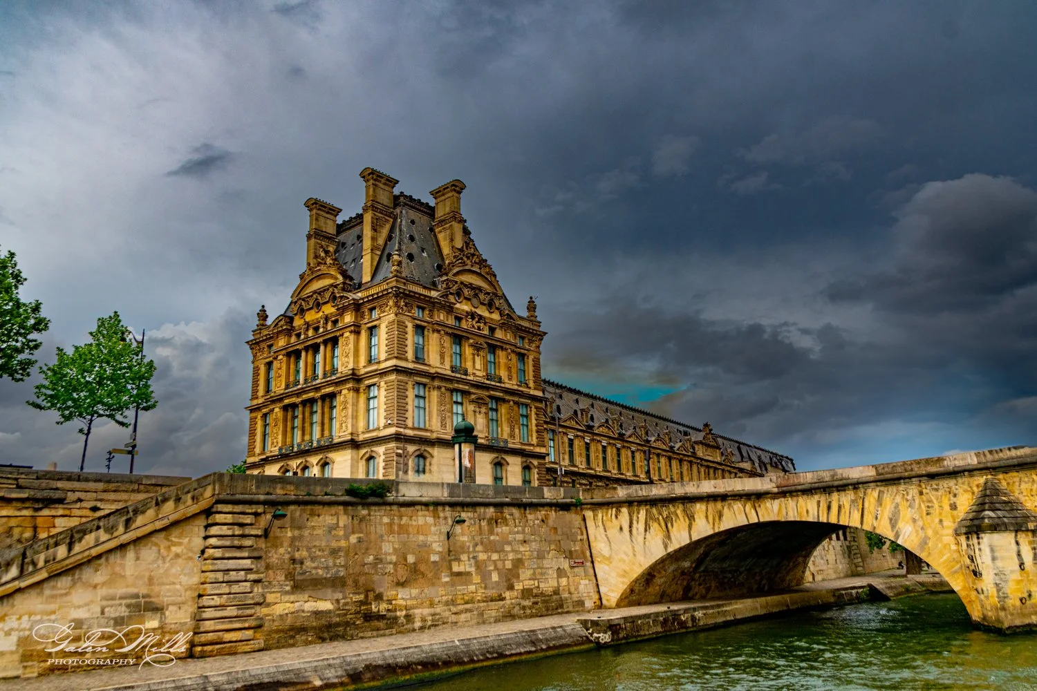Historic building with ornate architecture next to a stone bridge and river under cloudy sky.