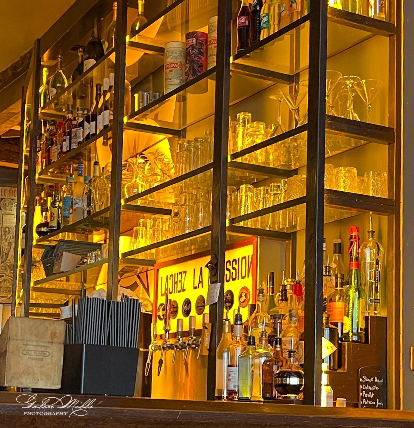 Bar shelves with various bottles, glasses, and a yellow illuminated sign with taps in a dimly lit setting.