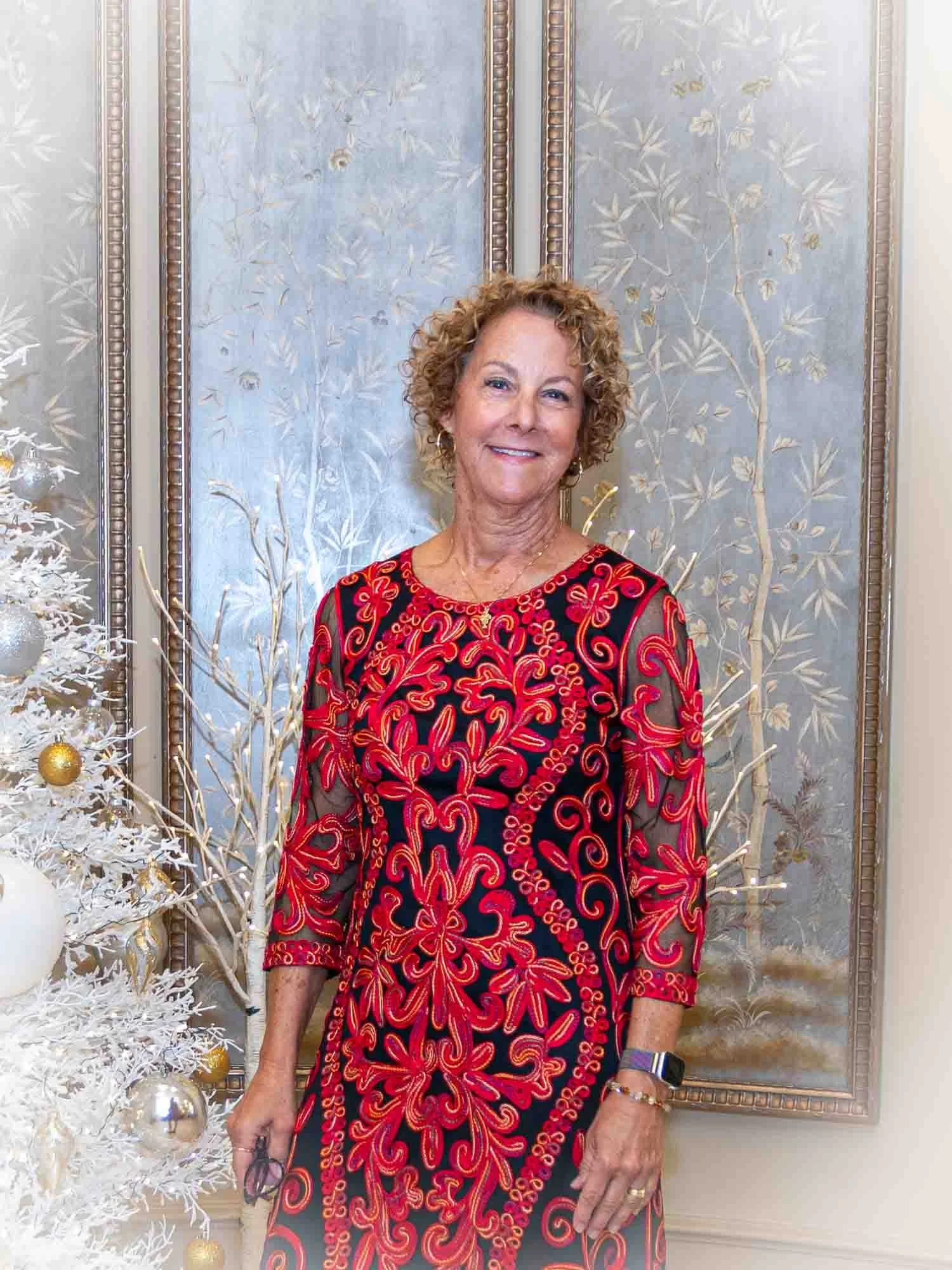 A woman in a red and black patterned dress stands in front of decorative panels and a white Christmas tree with ornaments.
