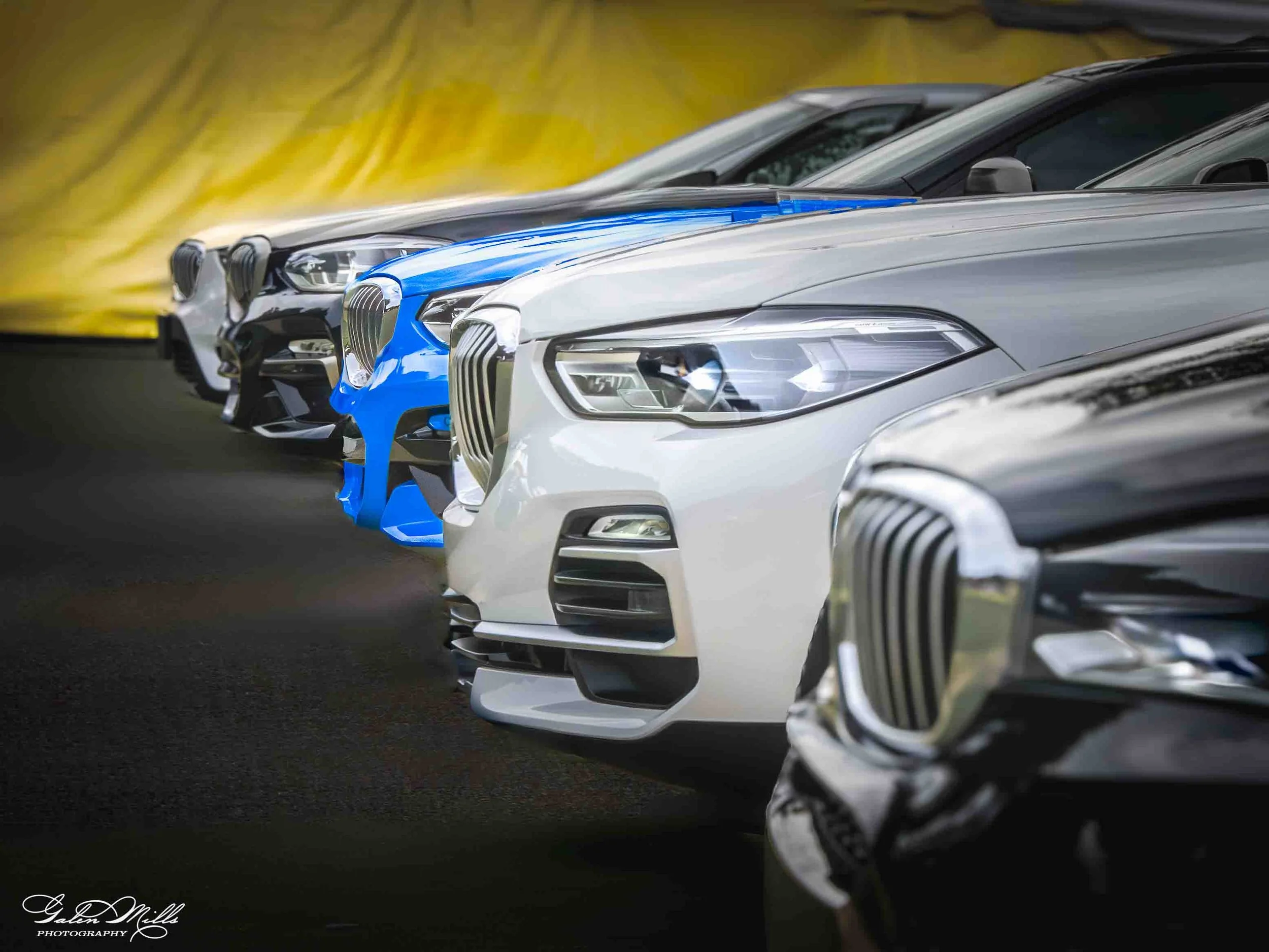 Close-up of several luxury BMW cars parked side by side, featuring modern headlights and grilles, with a yellow background.