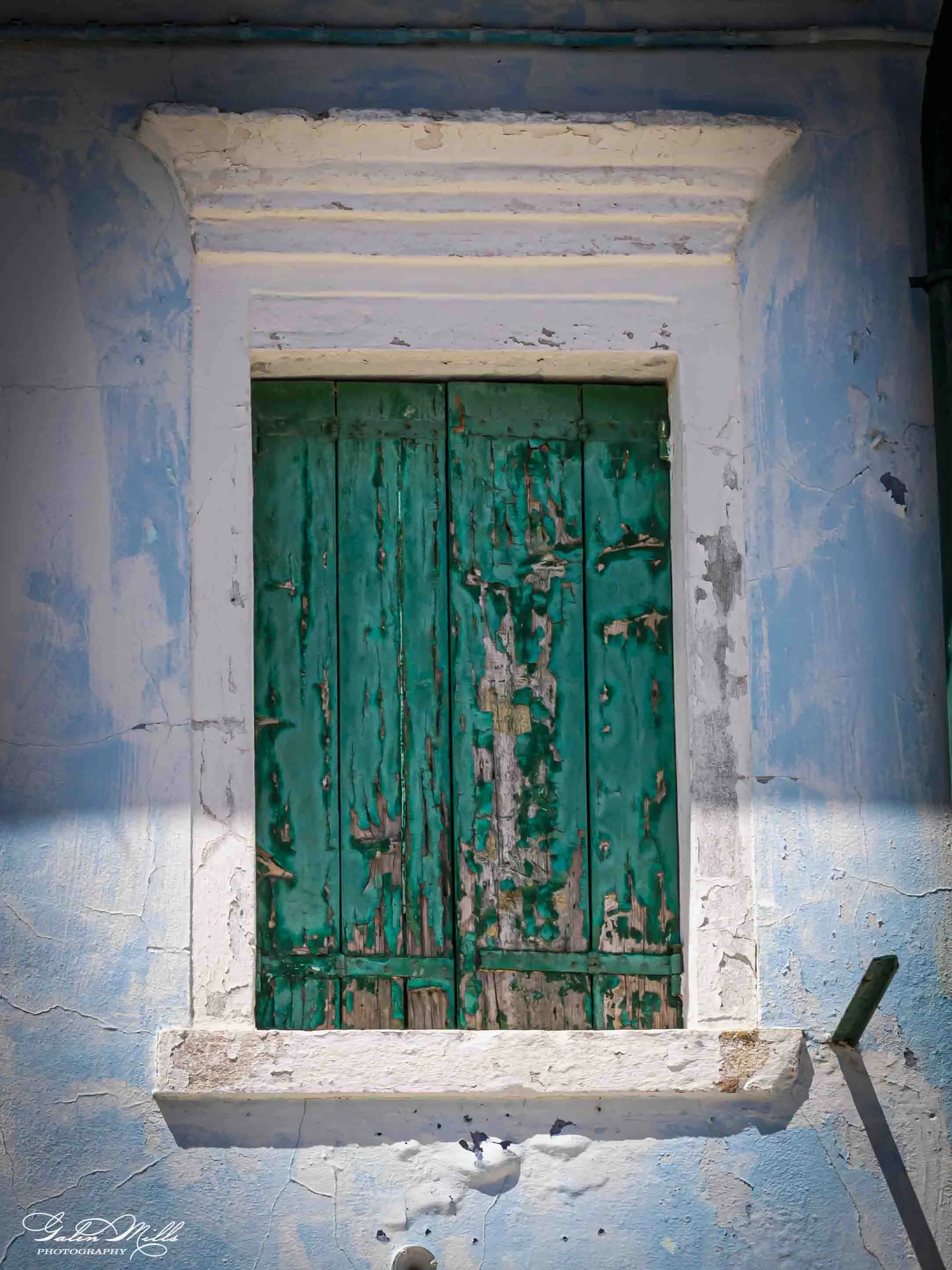 A weathered, closed green wooden door set in a white wall with peeling paint. The door is surrounded by a framed white trim and an arched top. Above the door is a decorative horizontal wooden panel with peeling paint. To the right of the door is a bl