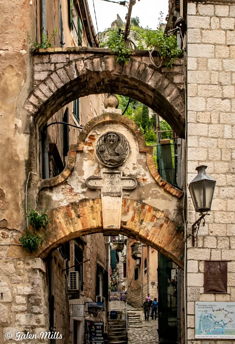 Old stone archway in a narrow street with historical inscription and weathered architecture, located in a European city. Small plants grow on top, and cobblestone pathway is visible below.