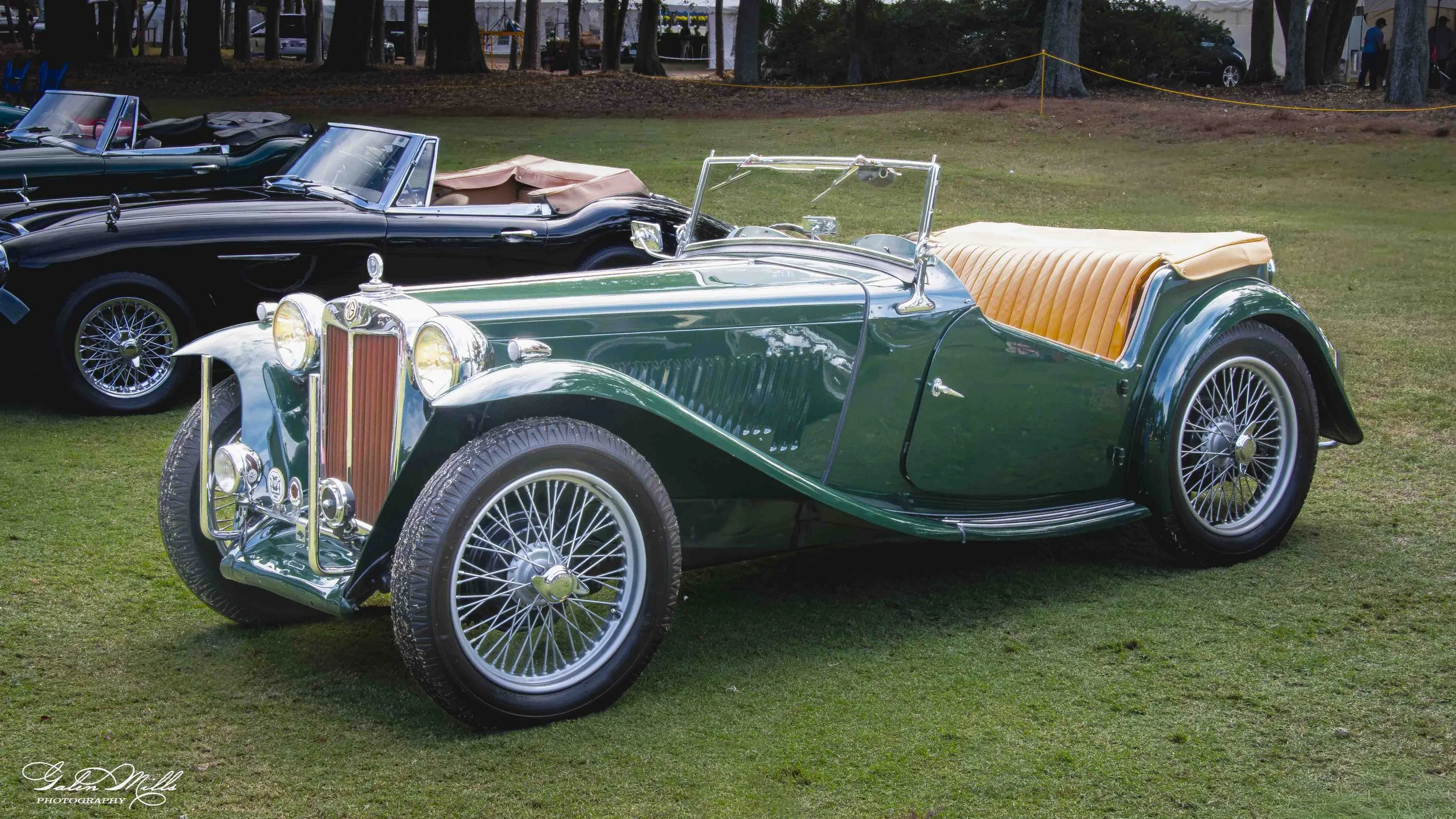 A vintage green convertible car with a tan interior parked on grass, alongside other classic cars, at an outdoor car show.
