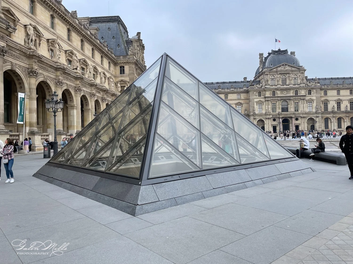 A glass pyramid structure at the Louvre Museum in Paris, with the historic museum building in the background, people walking around the plaza.
