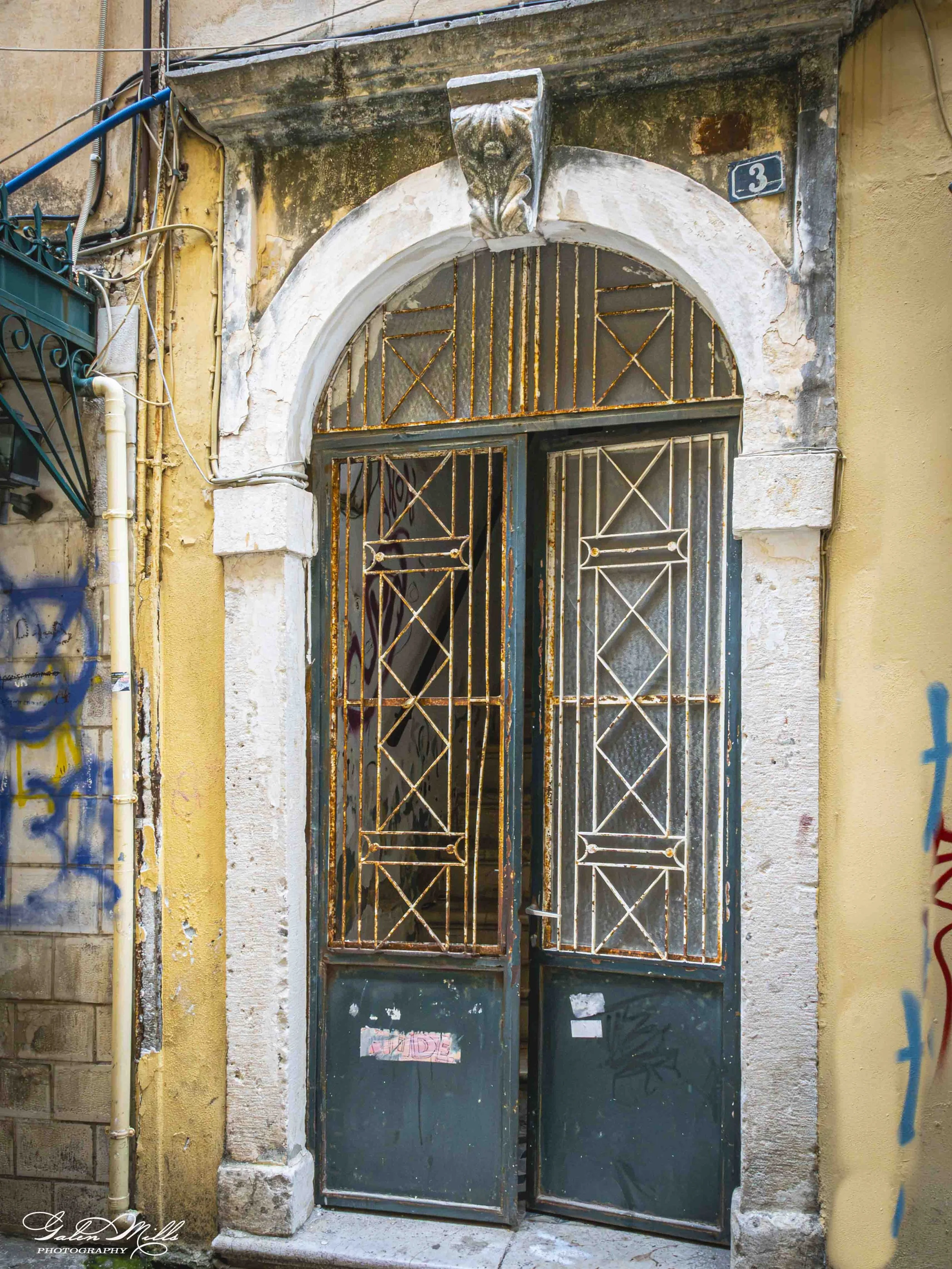 Old metal gate with rust and graffiti, set in a weathered stone and stucco wall. The arch above the gate is chipped and stained. The wall is yellow and beige, with visible cracks and peeling paint. A small blue house number plate reading '3' is affix