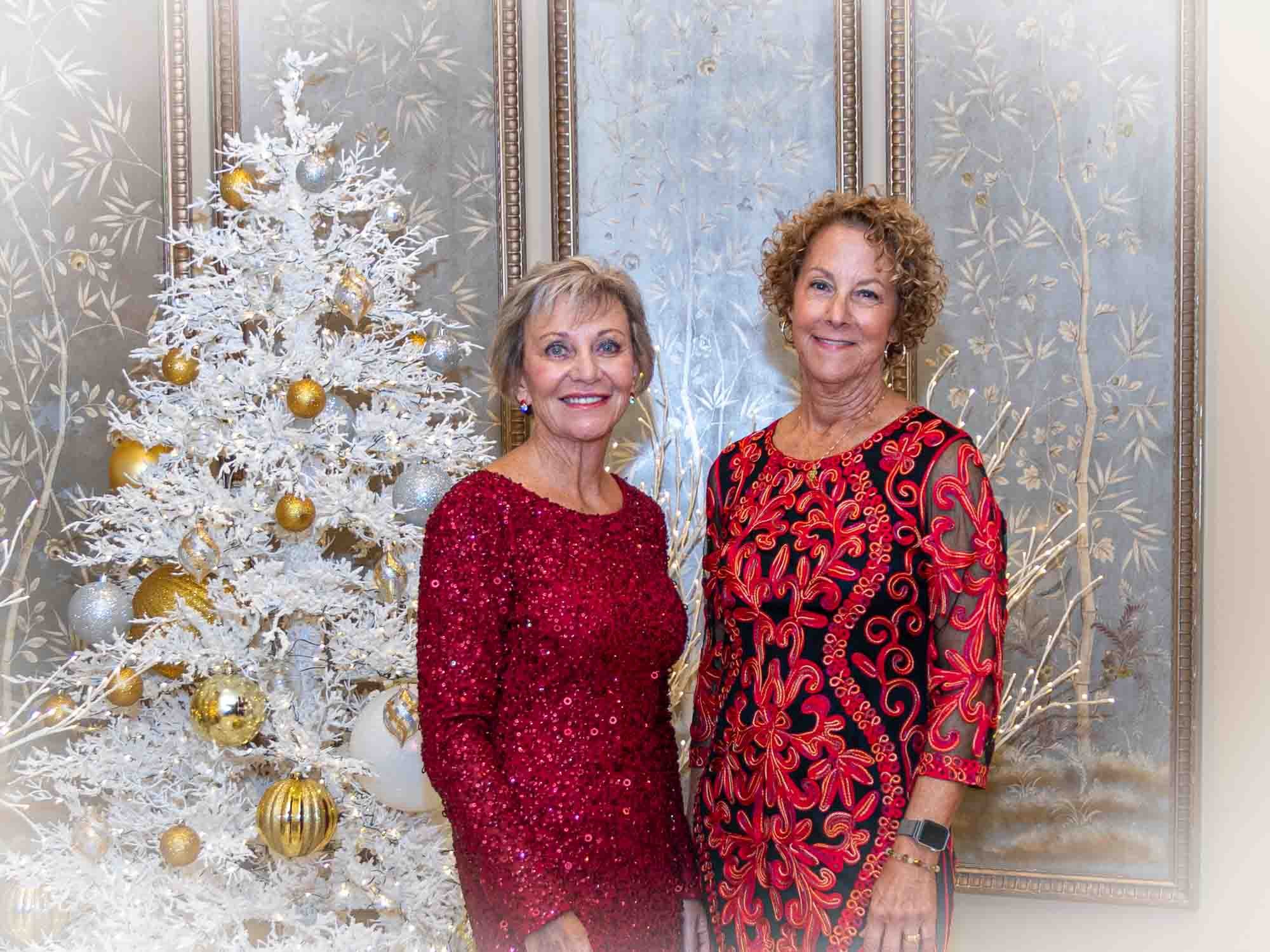 Two women in red dresses standing next to a decorated white Christmas tree.