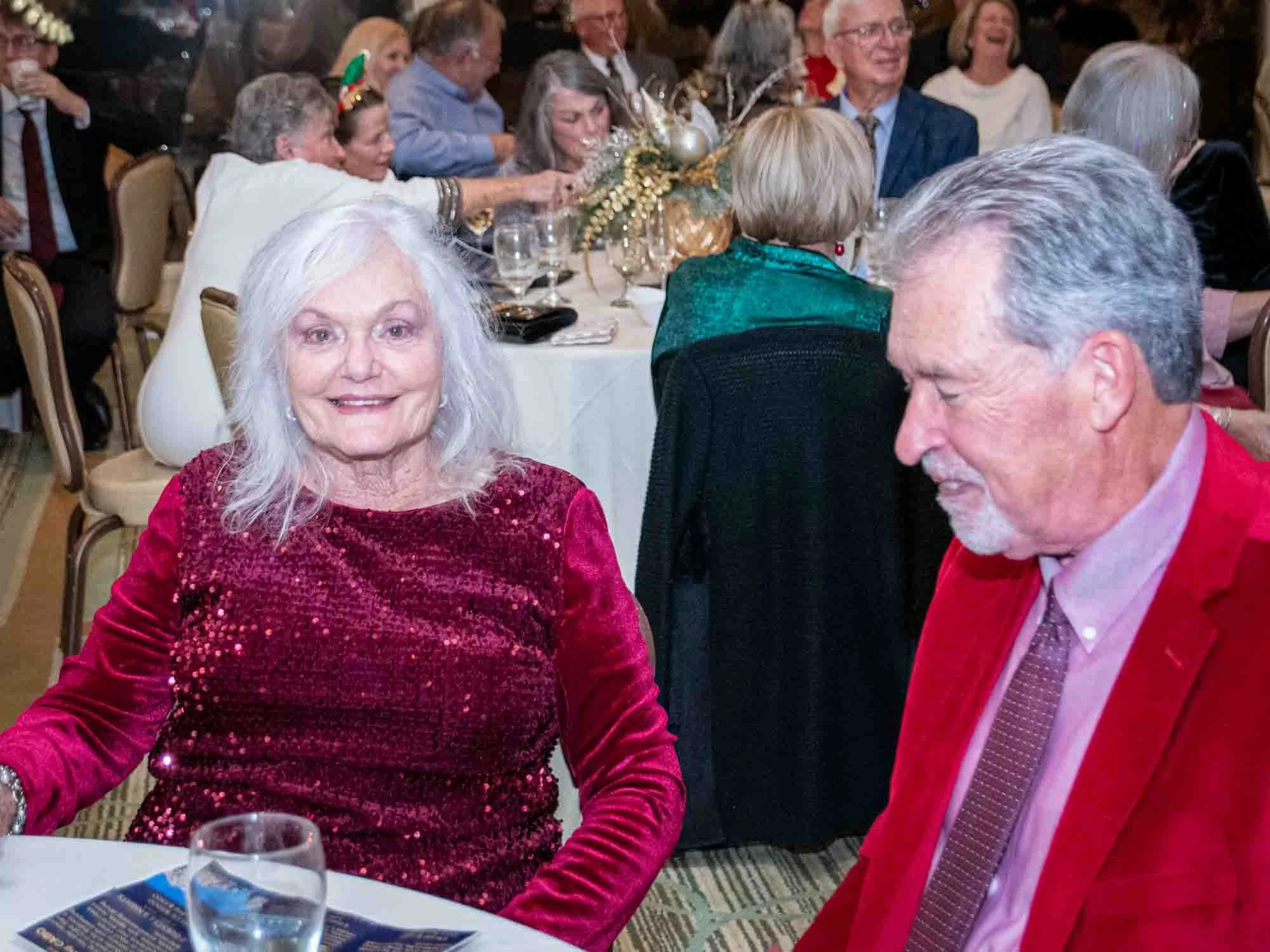 Elderly woman in sequined red dress sitting at a table with a man in a red blazer at a formal gathering.