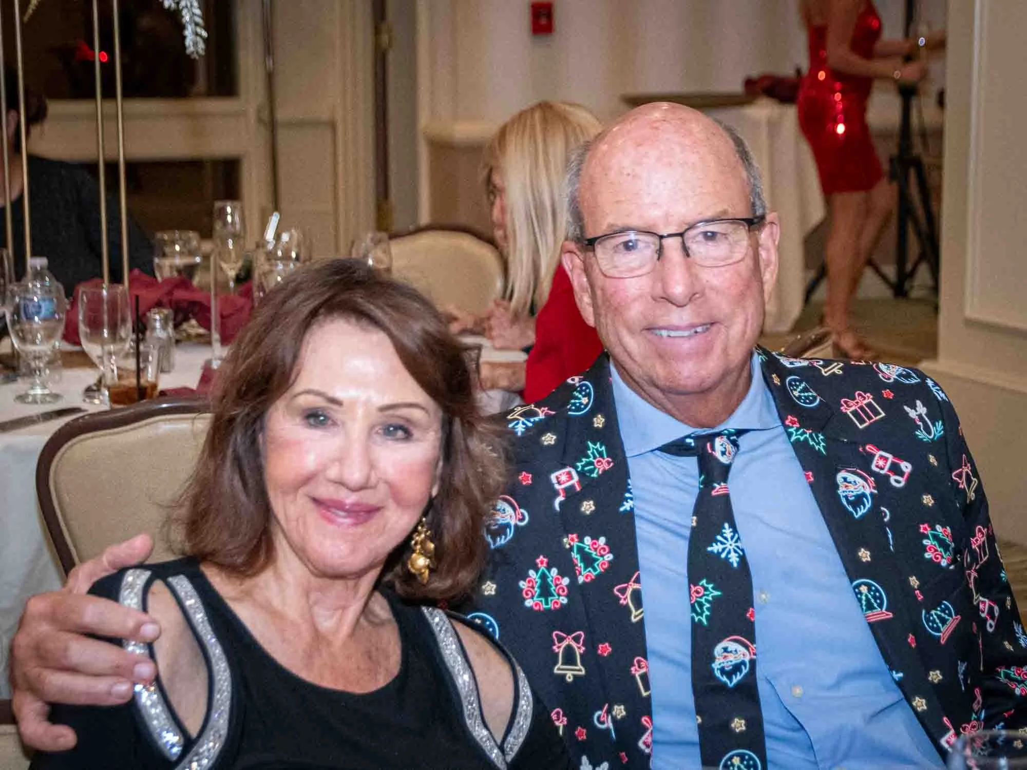 A couple sitting at a table during a festive event. The man is wearing glasses and a suit with a Christmas-themed pattern, and the woman is in a black dress with decorative sleeves. The background shows elegantly set tables and another person in a re