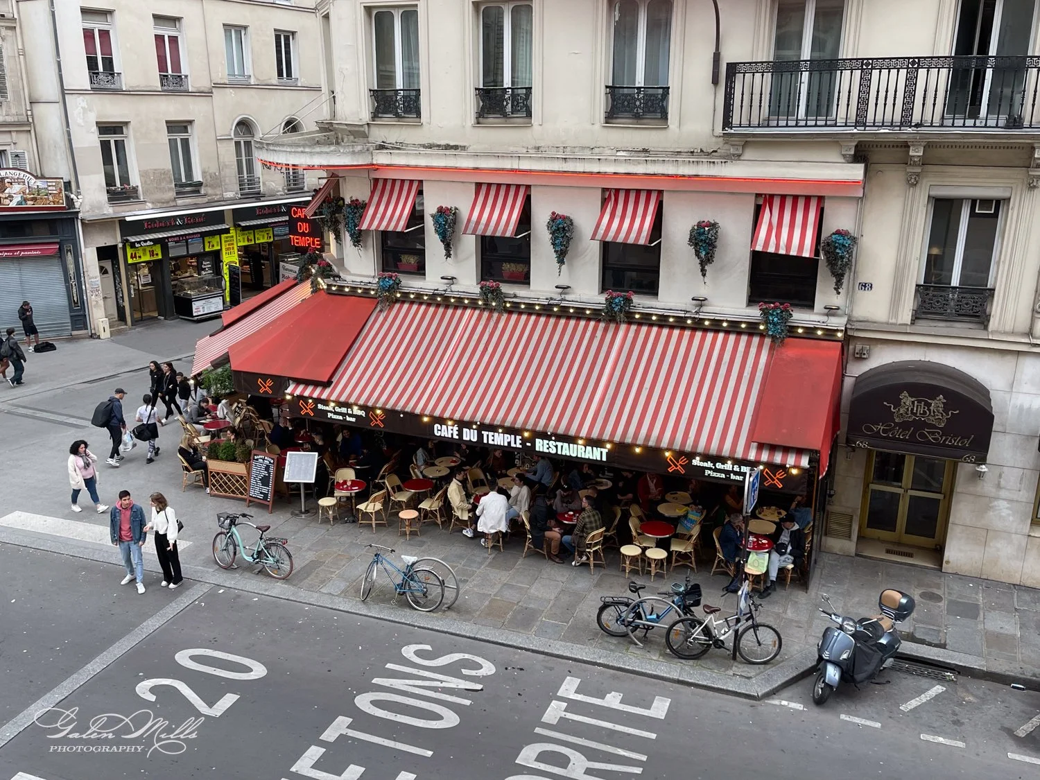 Street view of a Parisian café with striped red and white awnings, filled with people sitting at outdoor tables, located next to a hotel entrance. Bicycles are parked nearby on the sidewalk.