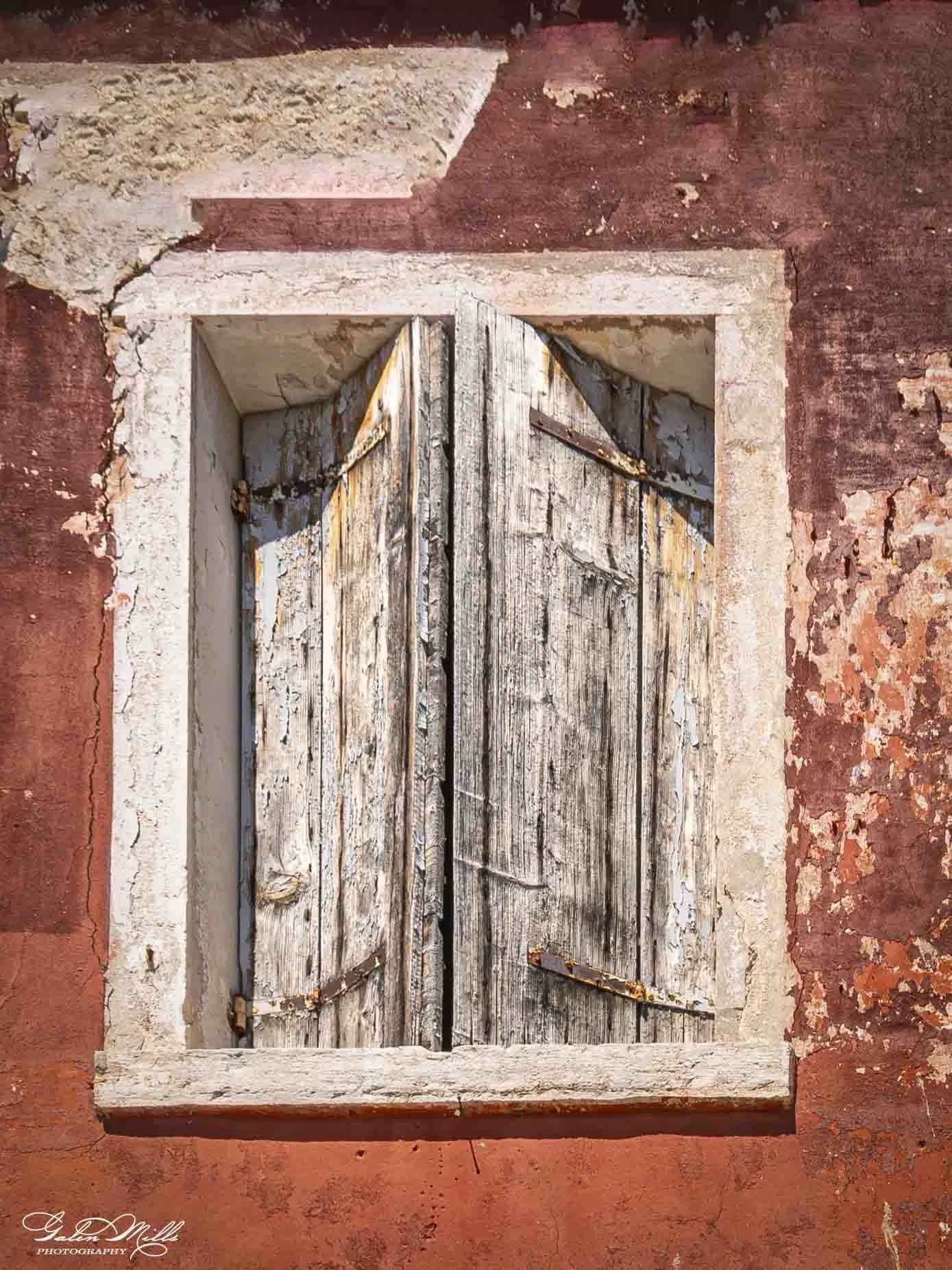 Old, weathered wooden window with peeling paint in a red stucco wall.