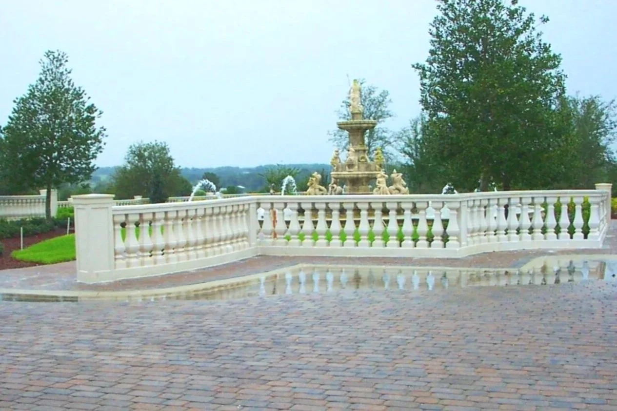 A decorative stone fountain with statues is surrounded by a white balustrade, situated on a cobblestone pavement, with trees and a cloudy sky in the background.