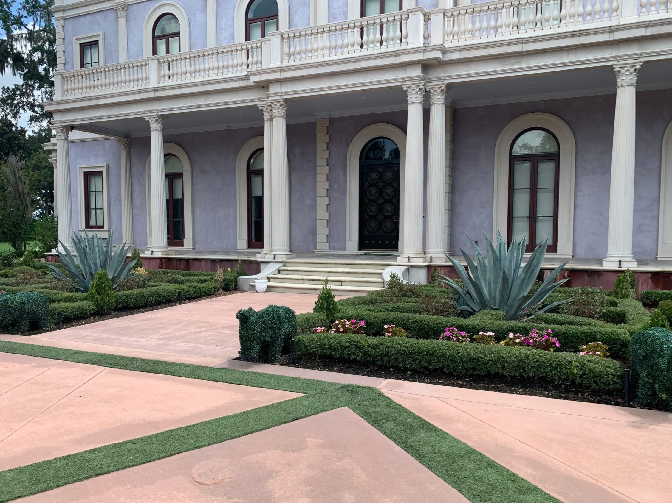 A large, elegant house with a light purple exterior, white precast columns supporting a balcony, and a black front door. The house is surrounded by well-maintained garden with various shrubs, flowers, and Agave plants, with a pinkish concrete pathway