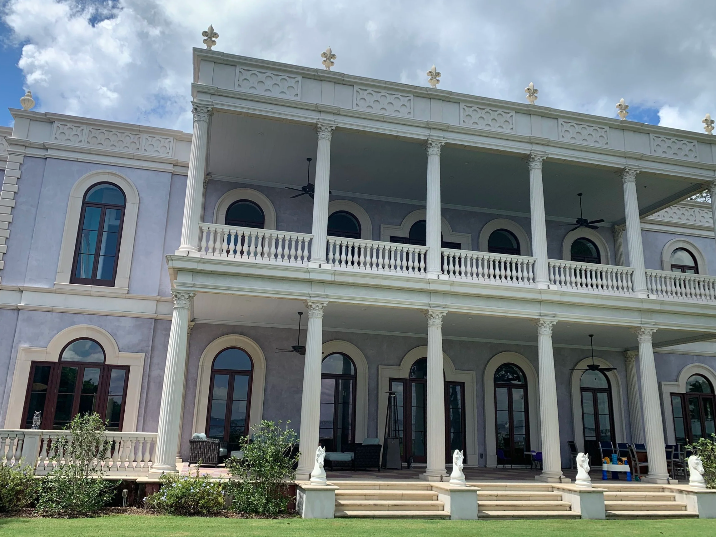 A large, white, multi-story house with classical architectural features, including columns and balustrades, with a spacious balcony and multiple windows, set in a well-maintained lawn with some greenery and small trees.