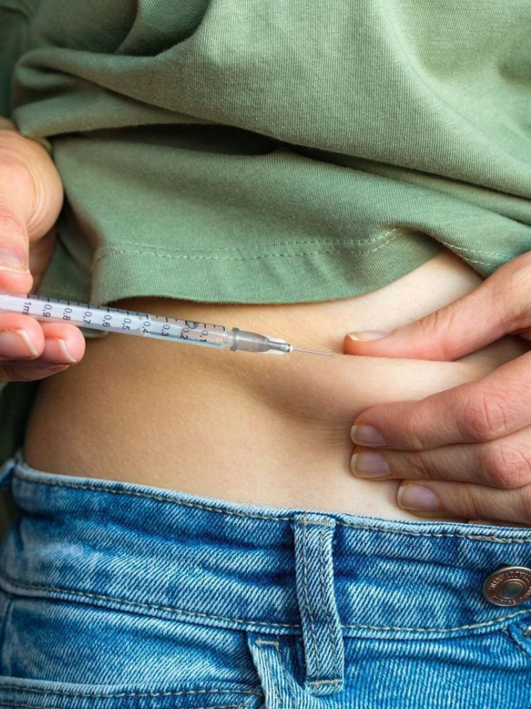 Person administering an injection into their abdomen, wearing a green shirt and blue jeans.
