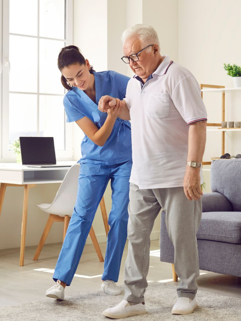 A healthcare worker assisting an elderly man with physical therapy exercises in a bright, modern living room.