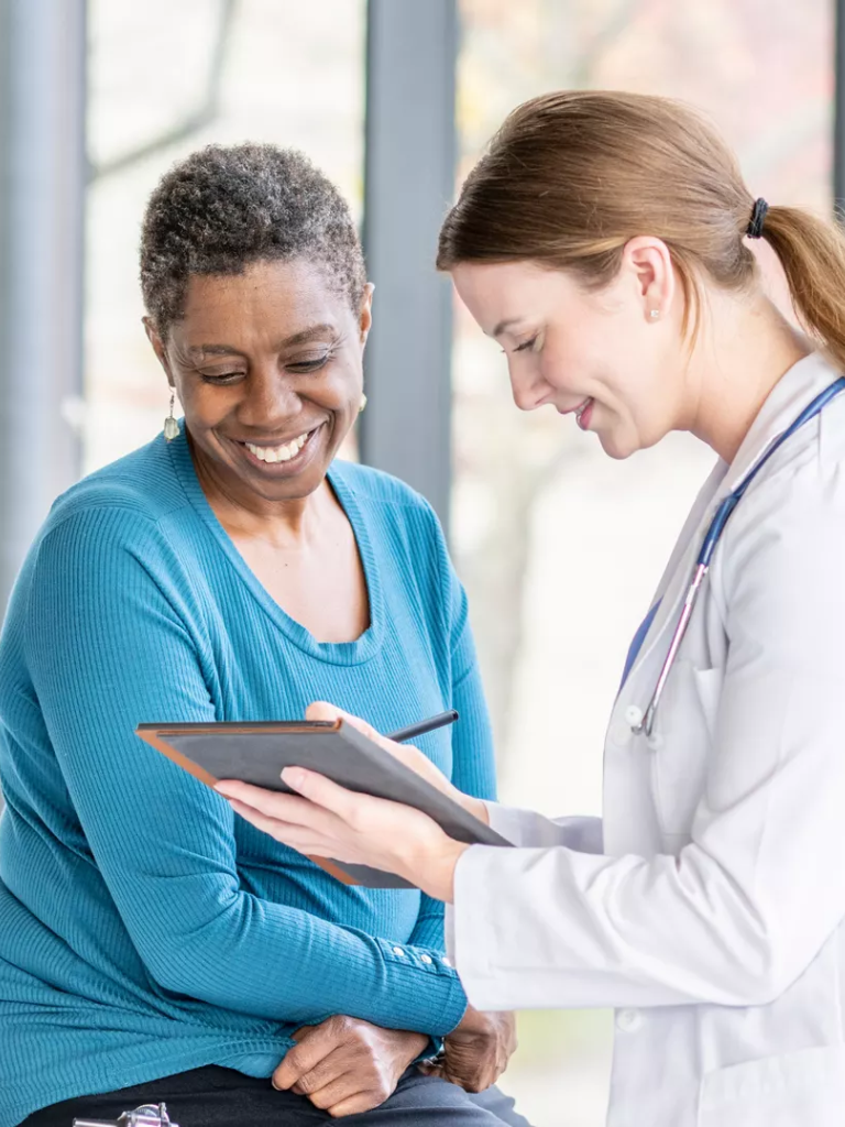 A female doctor and an older woman sharing a smile as the doctor shows her something on a clipboard in a medical setting.