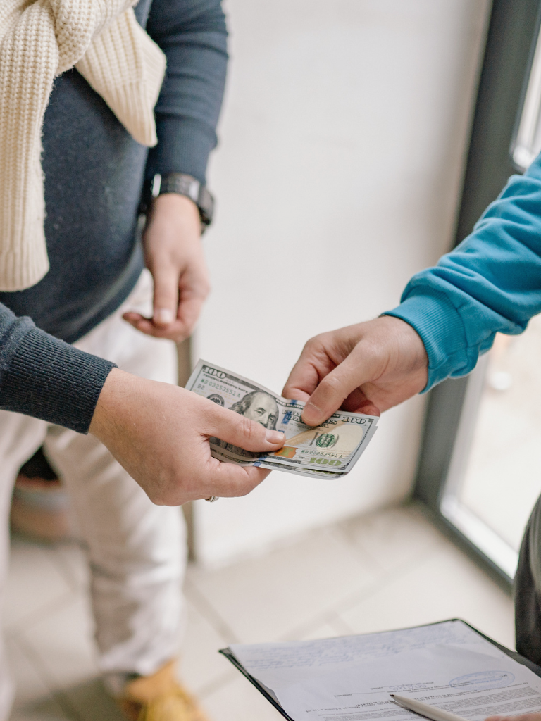 Person in a black sweater hands over money to another person in a blue sweater, near a door, with a clipboard on a table.