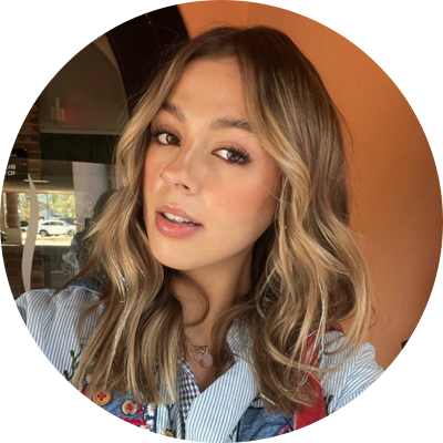 A young woman with wavy blonde hair and makeup, posing indoors with warm lighting.