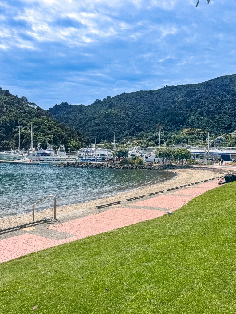 A scenic coastal area with a grassy lawn, a paved walkway, and benches along the shoreline. There are boats docked at a harbor against a backdrop of green mountains and a partly cloudy sky.