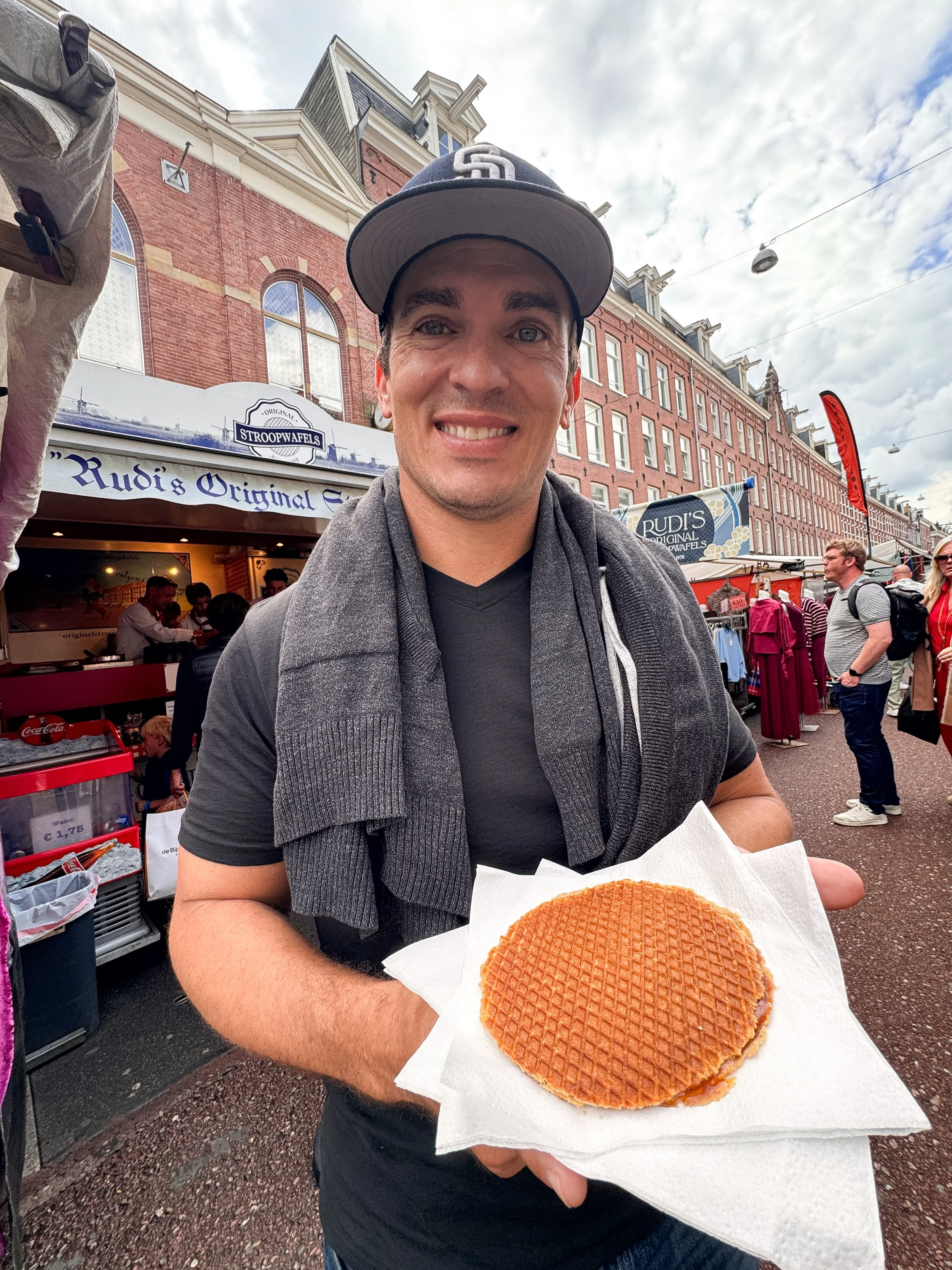 A smiling man wearing a black cap, grey scarf, and a black shirt, holding a waffle on a paper towel at an outdoor market with brick buildings and people in the background.
