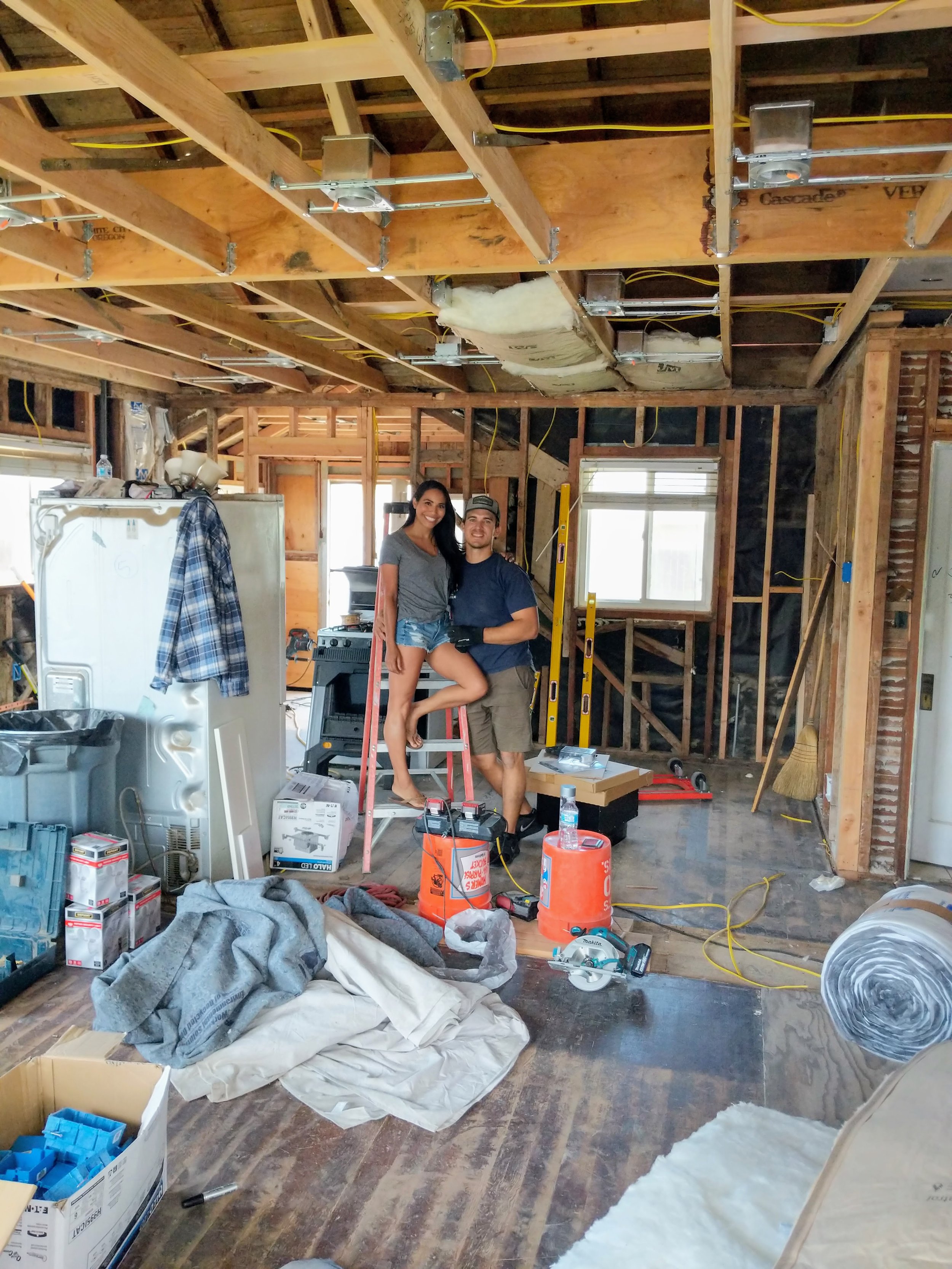Two people standing inside a house under renovation, with exposed wooden studs and ceiling, construction tools, and supplies scattered on the floor.