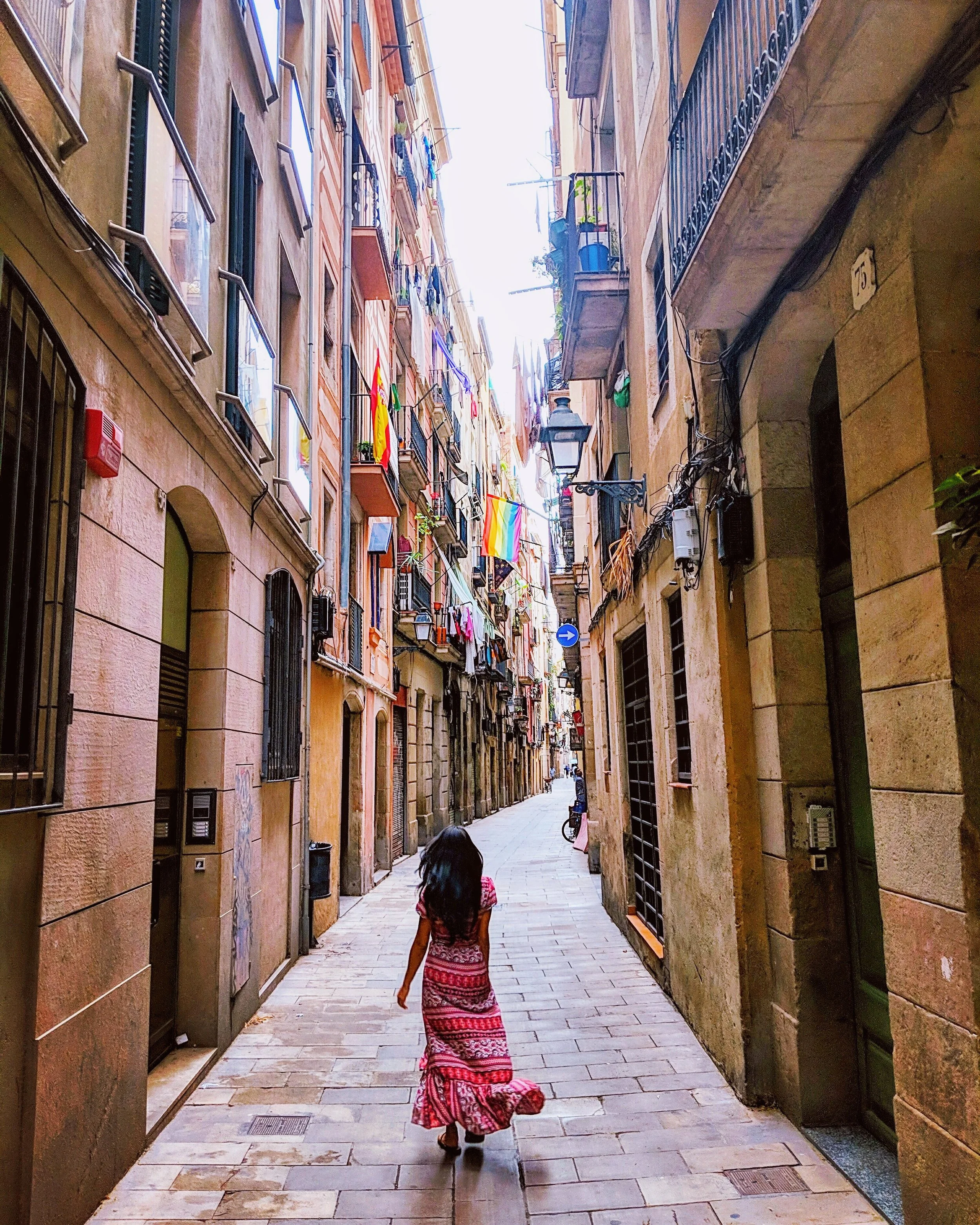 A girl in a long pink patterned dress walking down a narrow alleyway lined with multi-story buildings with balconies and windows. Clothes and flags are hanging on the balconies, and there is a bicycle parked further down the alley. The sky is bright at the end of the alley.