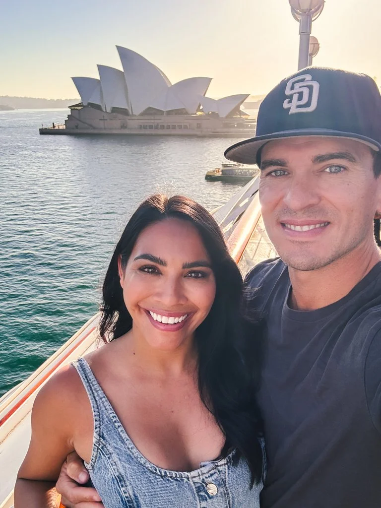A smiling couple taking a selfie on a boat with the Sydney Opera House in the background during sunset.